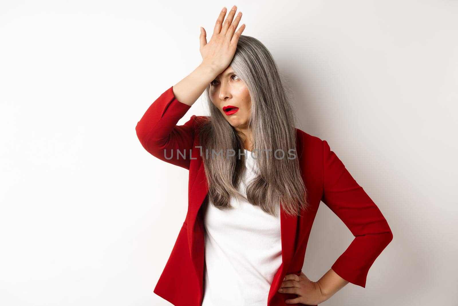 Annoyed asian office lady in red blazer roll eyes and facepalm, standing bothered and irritated over white background.