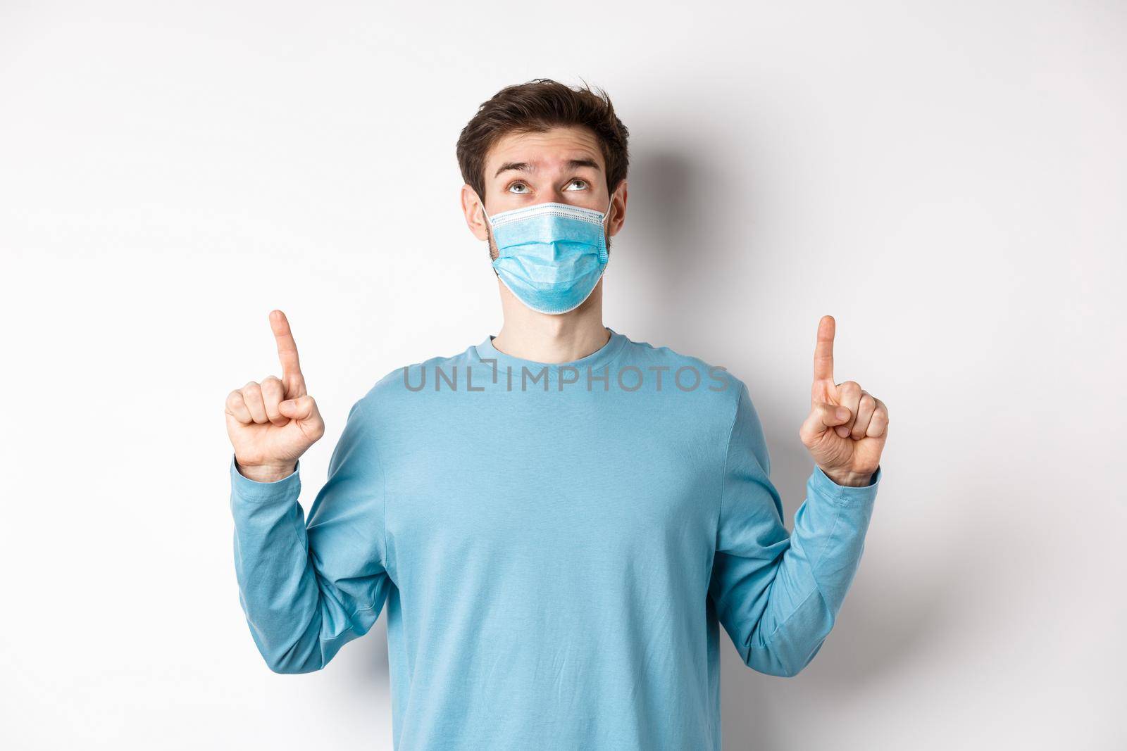 Coronavirus, health and quarantine concept. Curious guy reading banner on top, looking and pointing fingers up, standing in medical mask over white background by Benzoix