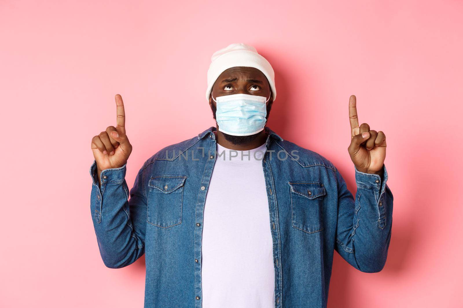 Coronavirus, lifestyle and global pandemic concept. Worried and anxious african-american man in face mask, pointing and looking up concerned, standing over pink background by Benzoix
