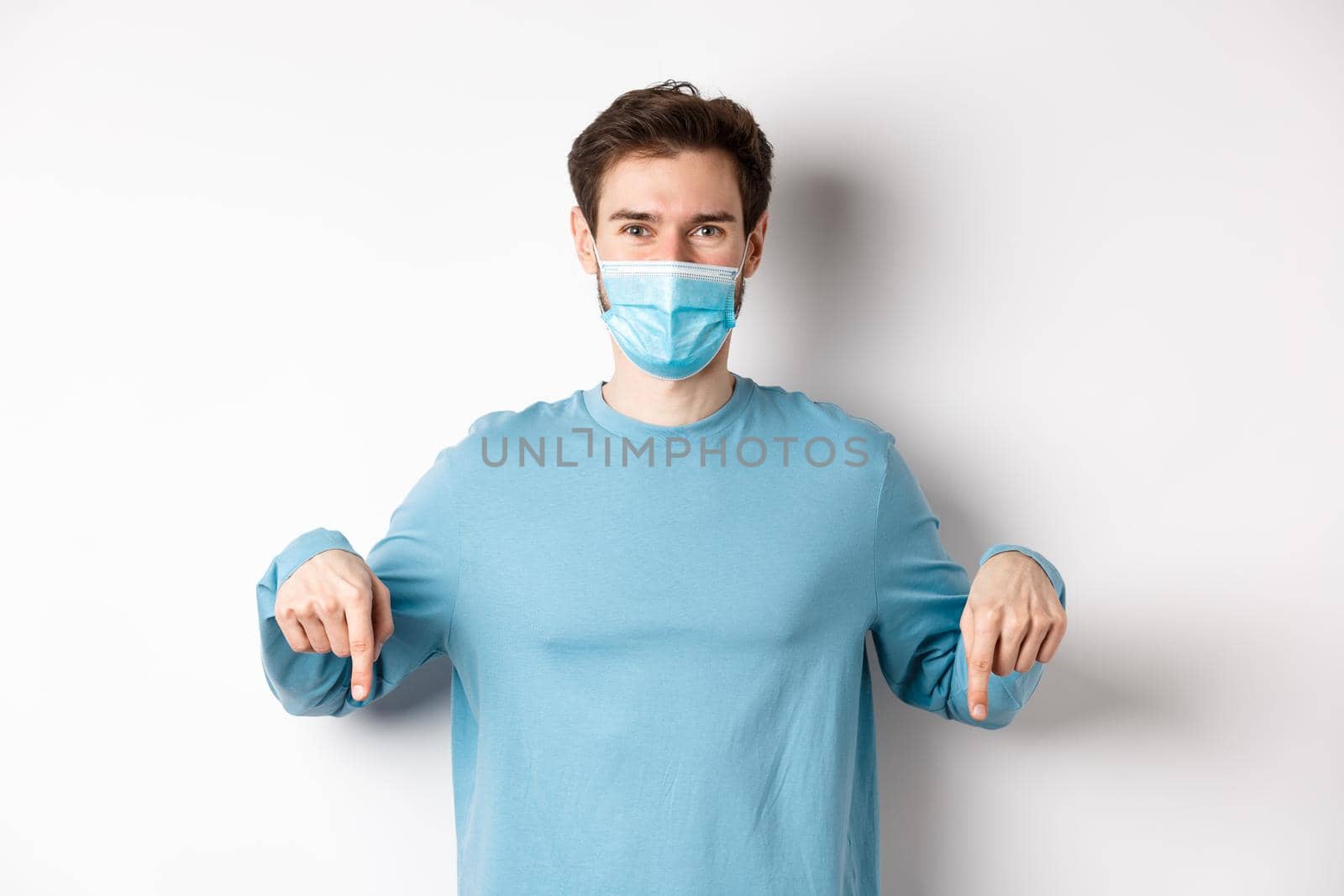 Covid-19, health and quarantine concept. Smiling caucasian man in face mask pointing fingers down, showing logo, standing over white background by Benzoix