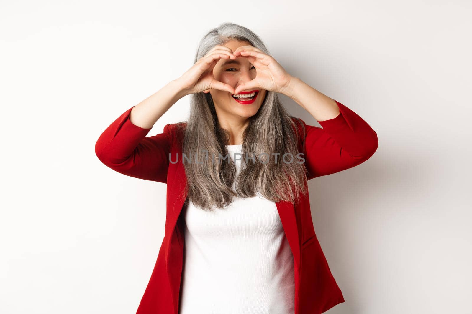 Lovely asian middle-aged woman with grey hair, wearing red blazer, showing heart sign and peeking throught it, I love you gesture, standing over white background.