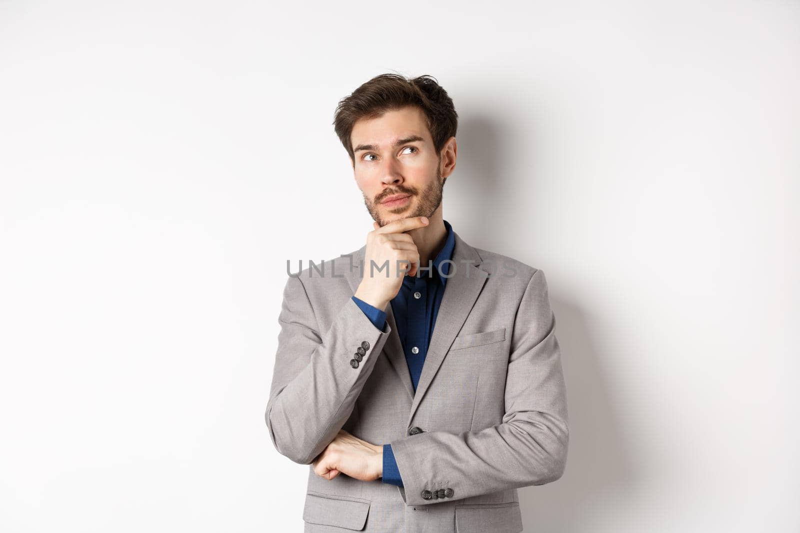 Pensive handsome man in suit looking up and thinking, making choice, pondering ideas, standing against white background.