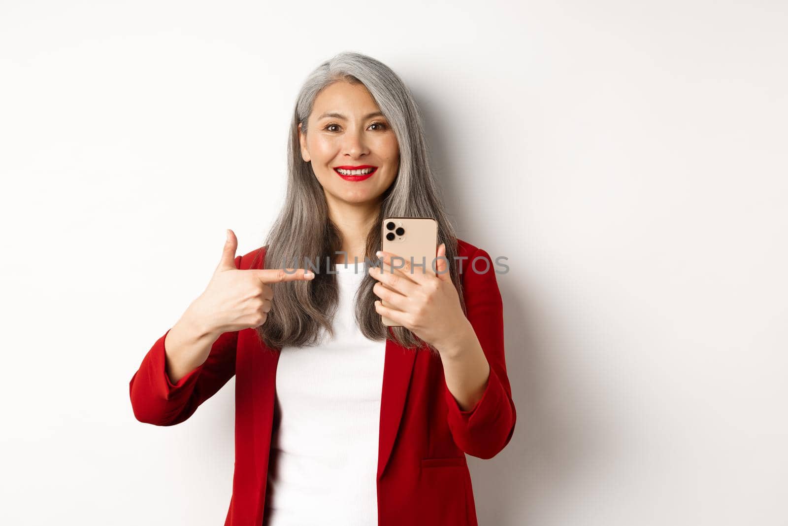 Asian elderly woman in elegant blazer showing smartphone, pointing finger at mobile phone and smiling, standing over white background.