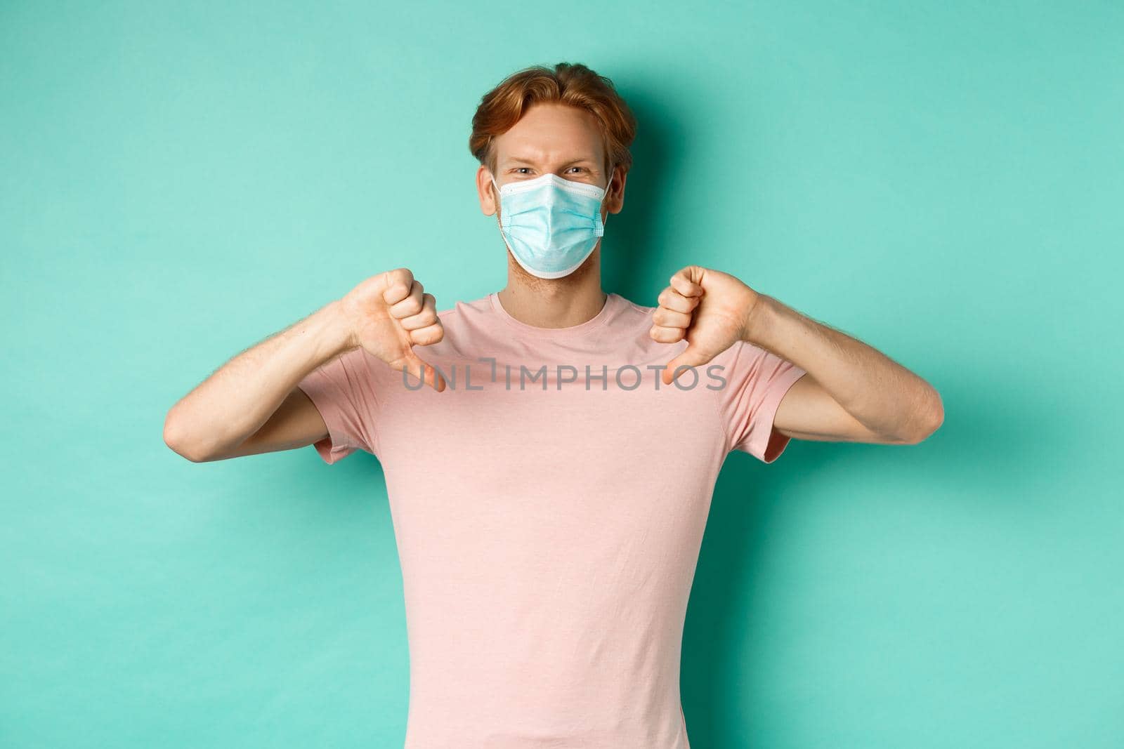 Covid-19, pandemic and lifestyle concept. Young man with red hair in face mask, showing thumbs down, dislike or disapprove something, standing over turquoise background.