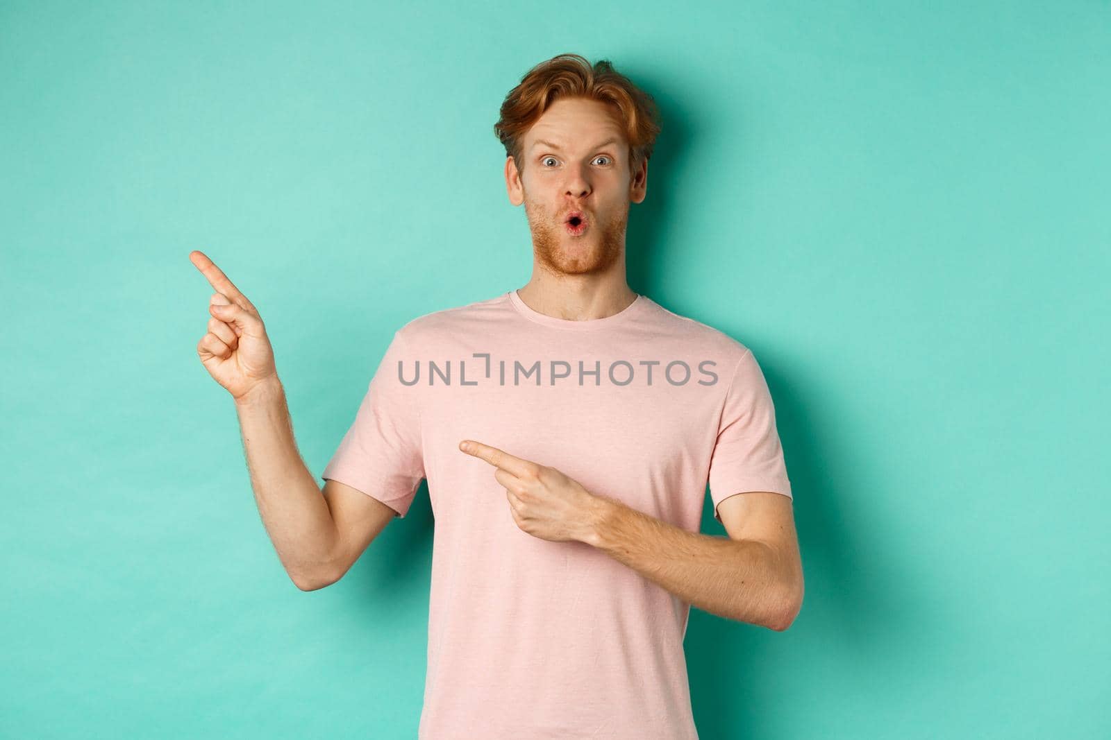 Amazed young man with red hair and beard, gasping amazed, pointing at upper left corner and showing promo, checking out advertisement, standing over turquoise background.