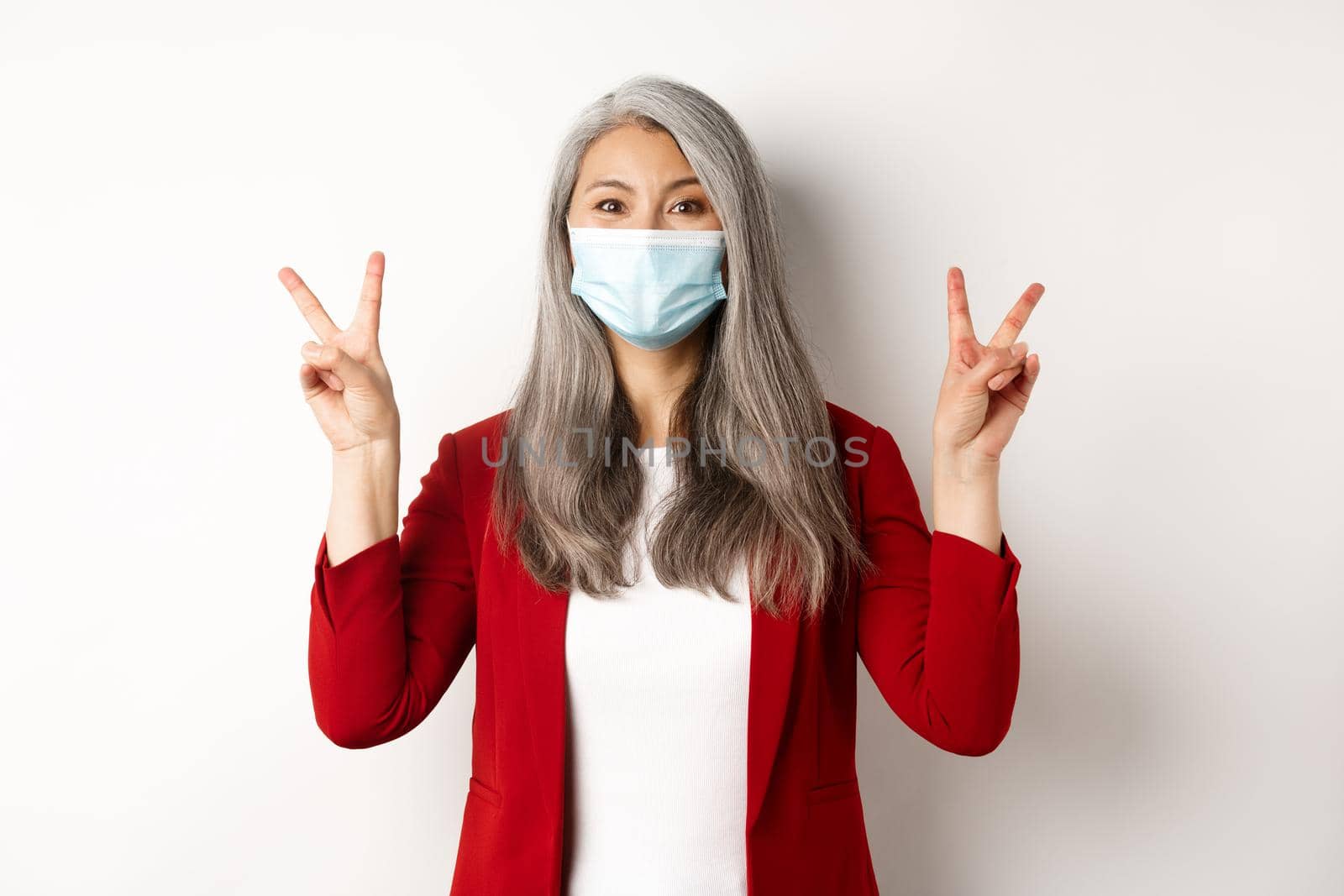 Coronavirus and business concept. Cheerful senior woman in face mask, showing victory or peace sign and smiling with eyes, white background.