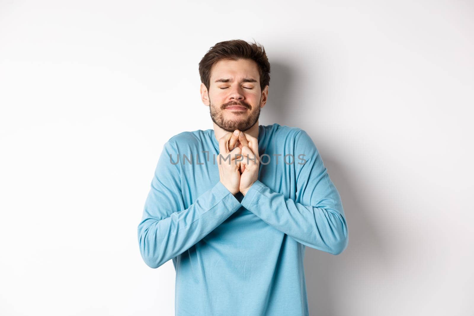 Hopeful caucasian guy in sweatshirt cross fingers for good luck. Young man making wish, praying with eyes closed and dreamy smile, standing over white background.