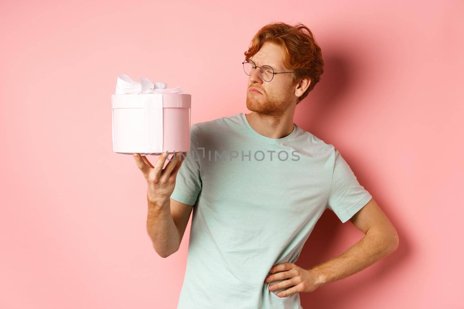 Love and holidays concept. Intrigued redhead guy looking puzzled at gift box, dont know what inside, standing over pink background by Benzoix