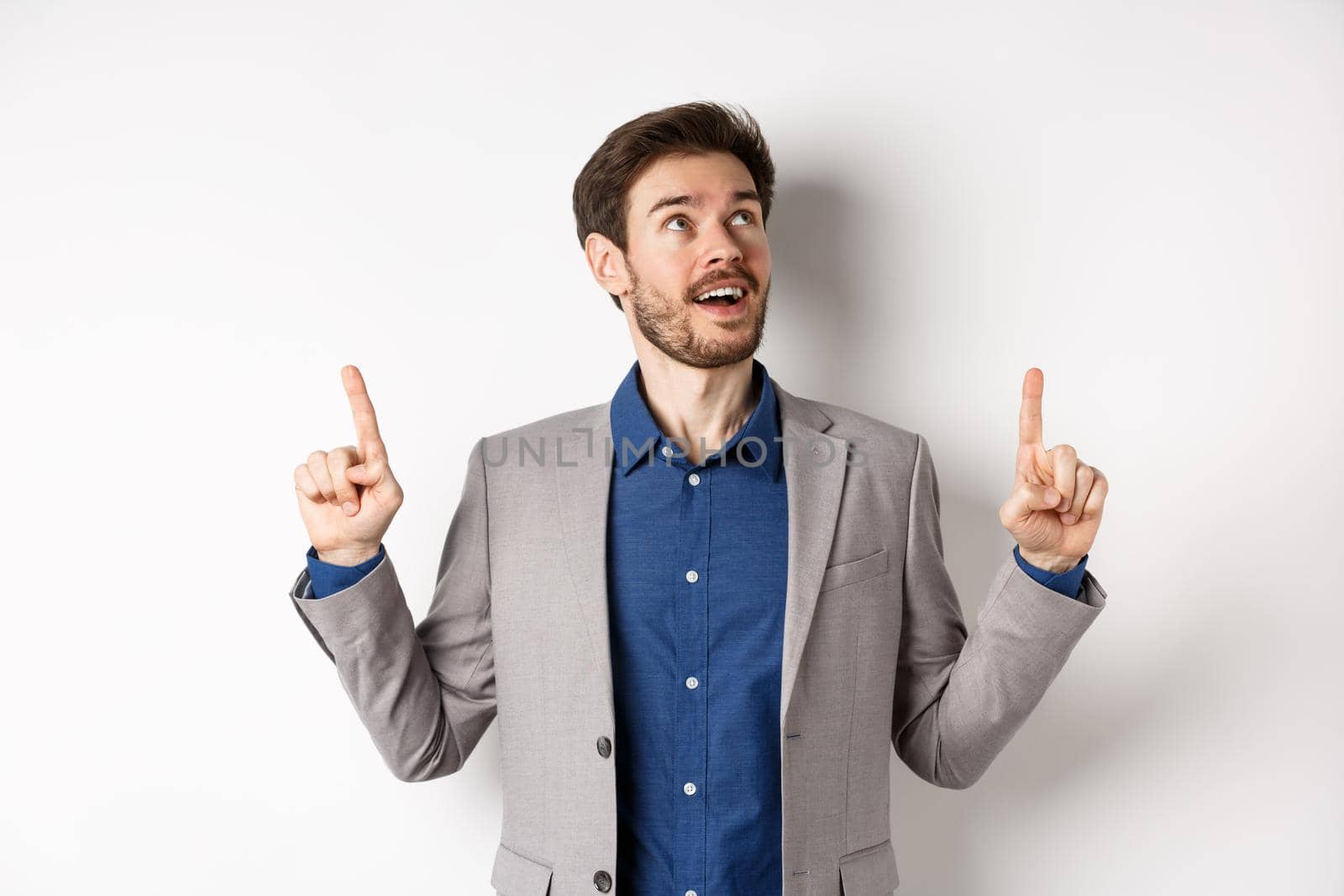 Excited handsome man with beard, wearing suit and looking amused up, pointing at top with admiration, standing against white background by Benzoix