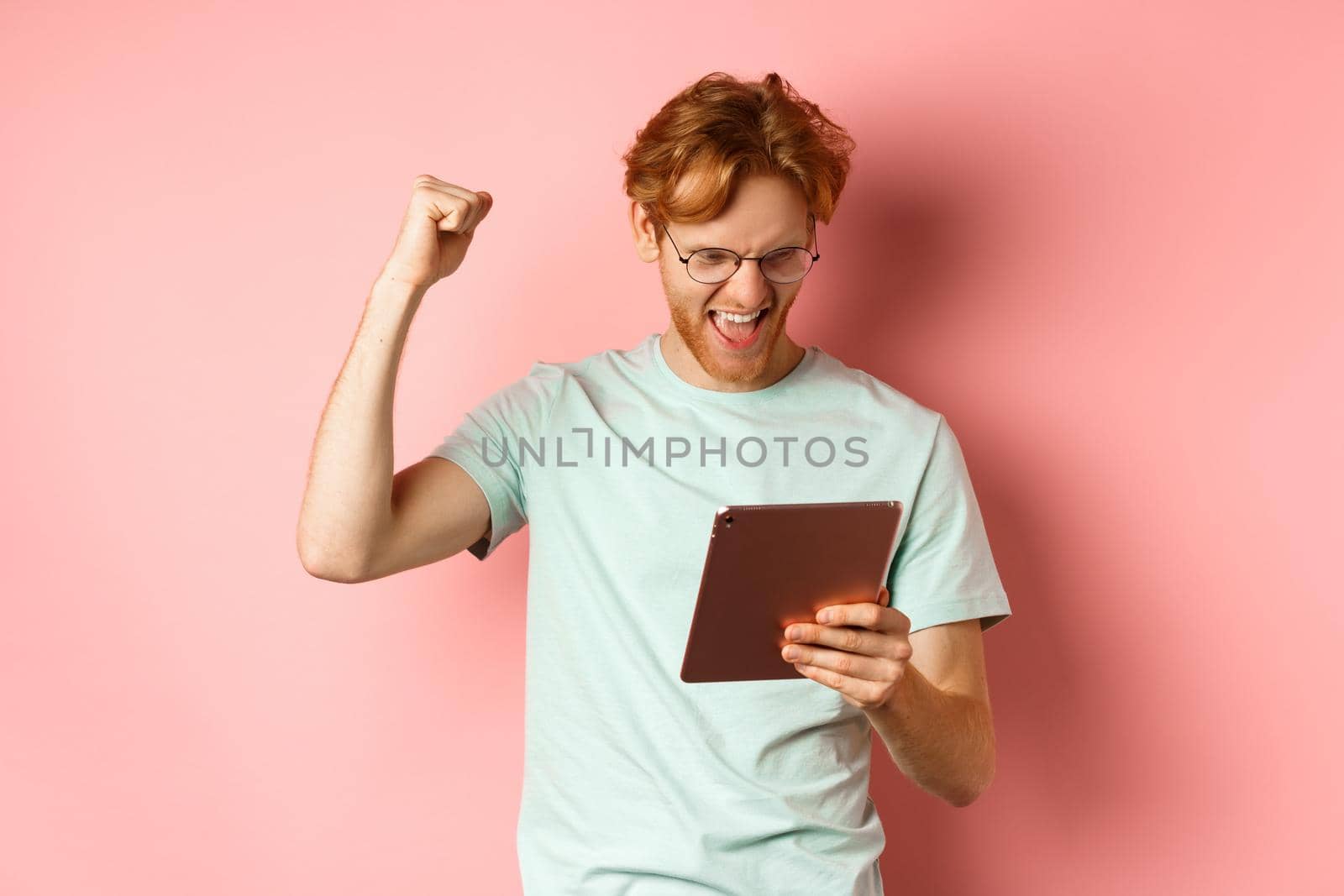Image of happy redhead man triumphing, winning online with digital tablet and rejoicing, standing over pink background.