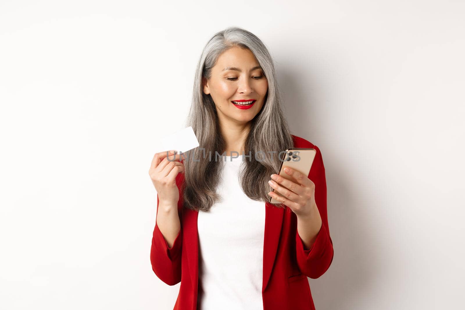 Asian senior woman in elegant outfit making payment online, holding plastic credit card and smartphone, white background.