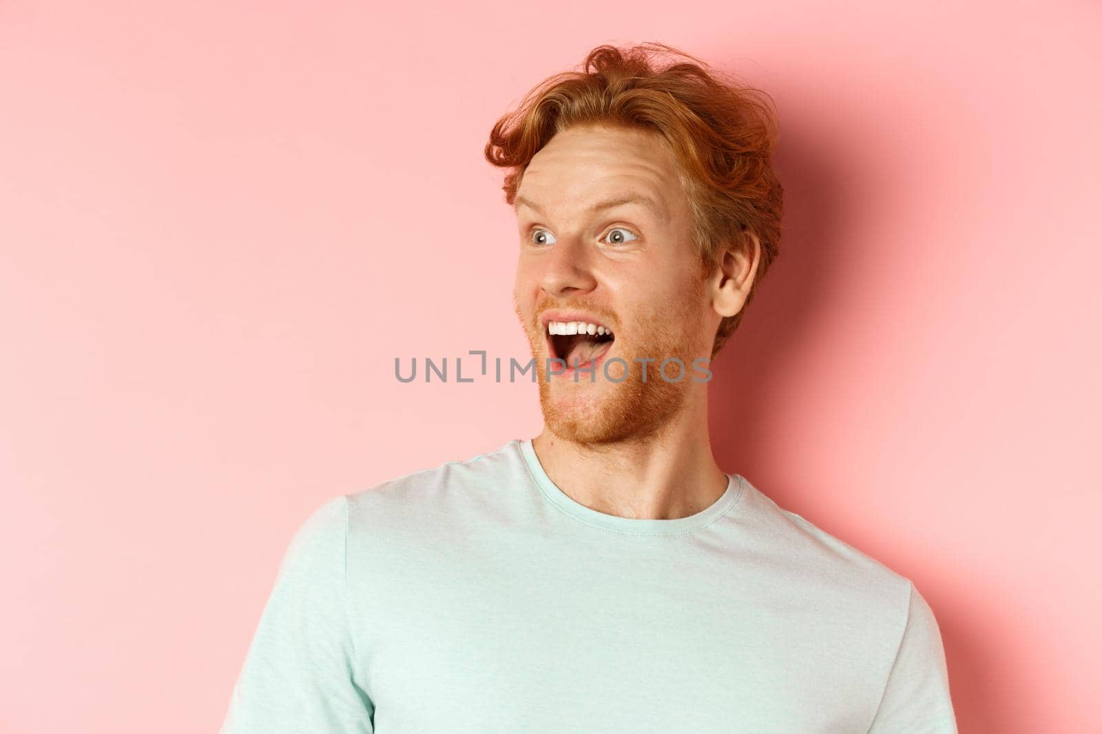 Close up of happy and fascinated young man with red hair checking out promo deal, looking left with amazed smile, standing over pink background.