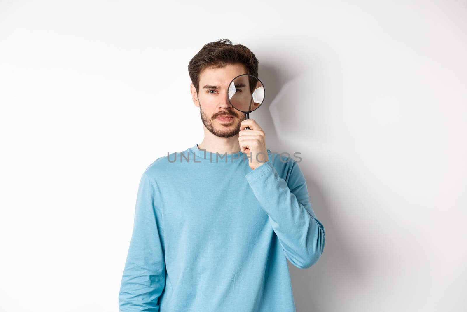 Man searching for something, looking through magnifying glass at camera, standing on white background.