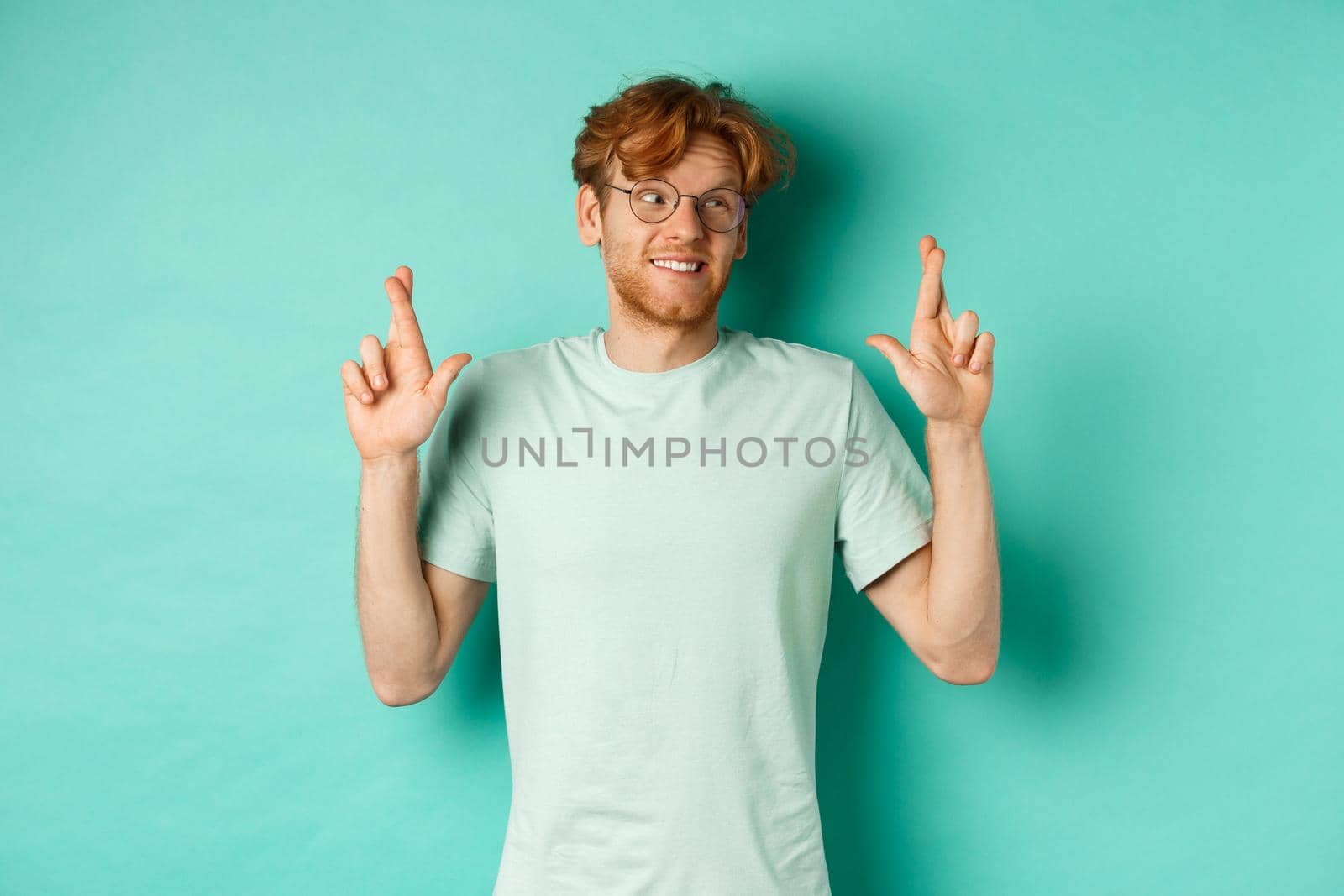 Hopeful redhead man in glasses making a wish, cross fingers and looking right at copy space, dreaming about something or making wish, standing over turquoise background by Benzoix