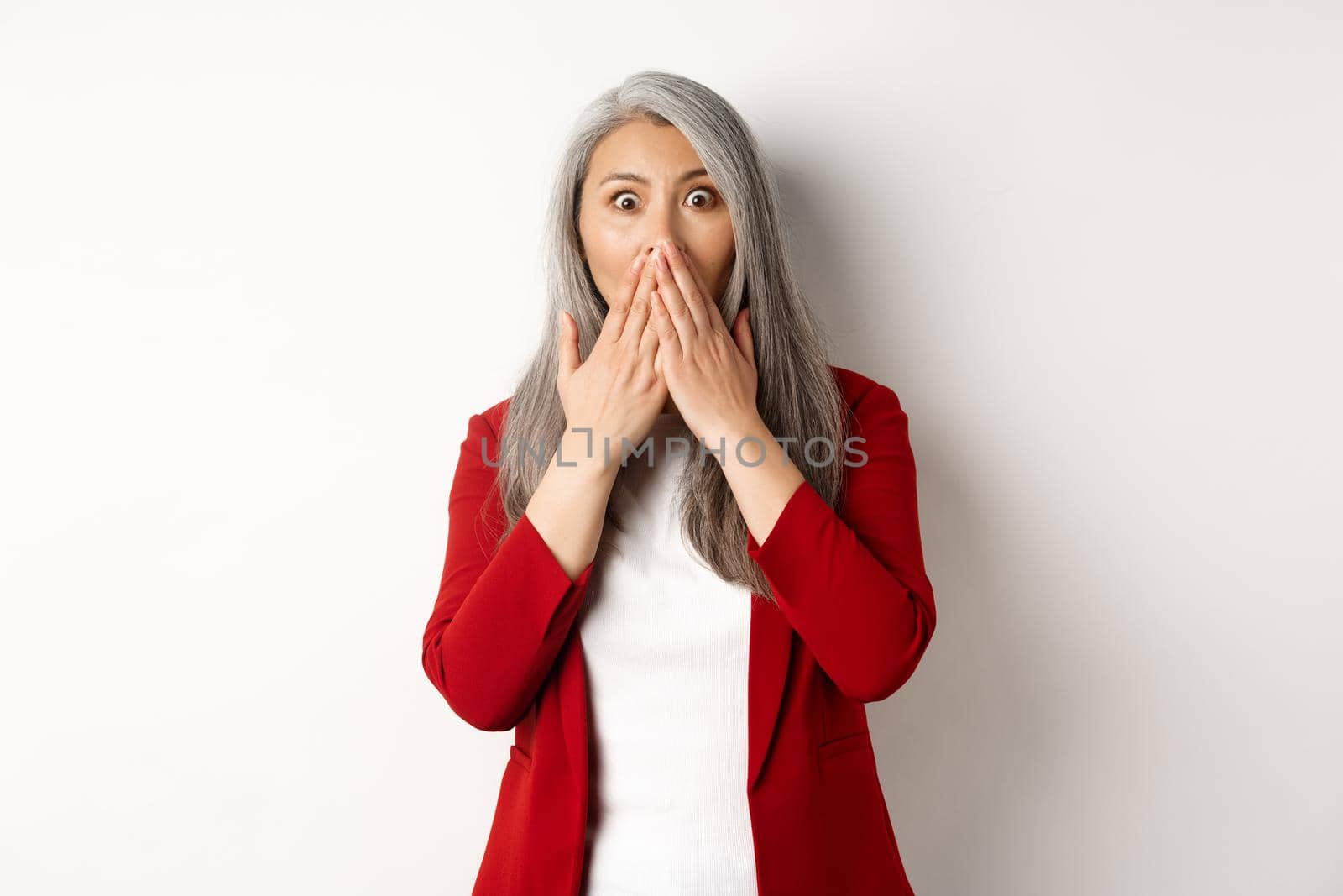 Shocked asian senior lady gasping, cover mouth from amazement, hear gossip, standing over white background by Benzoix