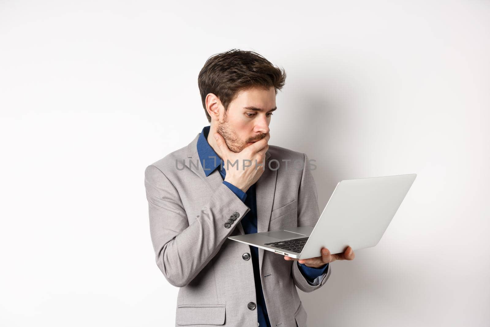 Shocked office worker in suit looking worried at laptop screen, having trouble at work, standing on white background.