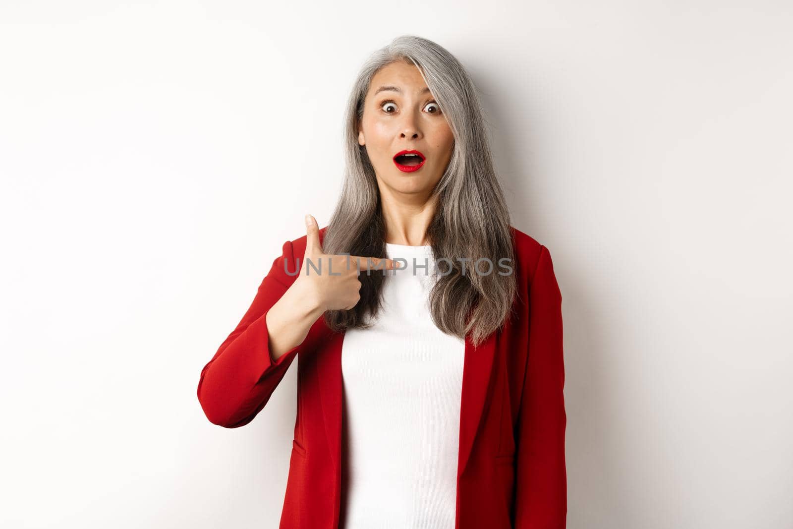 Surprised asian woman with grey hair, pointing at herself and gasping confused, standing over white background by Benzoix