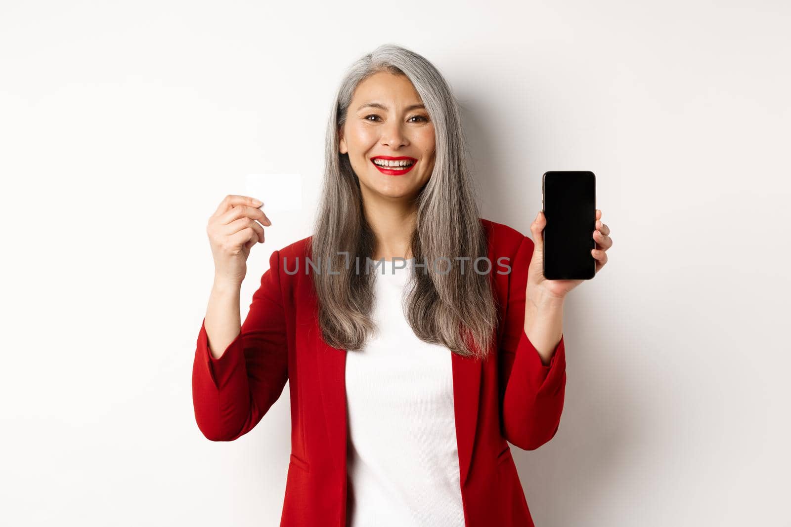 Senior asian businesswoman showing plastic credit card and blank smartphone screen, smiling at camera, white background.