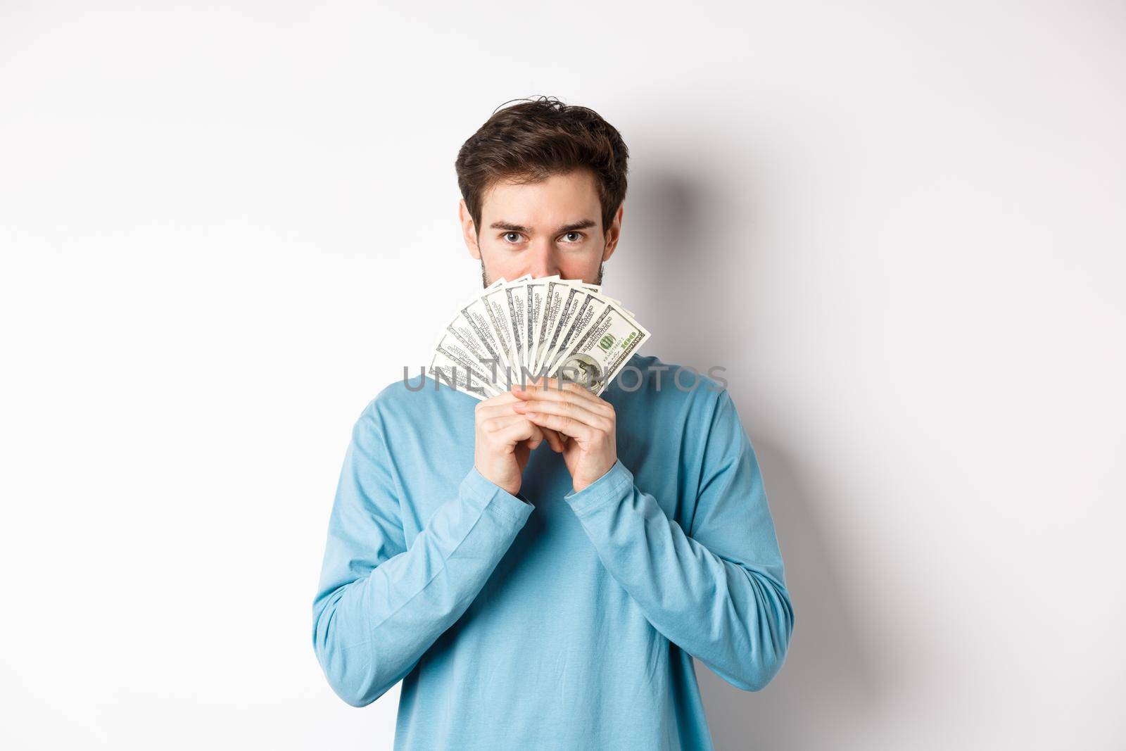 Finance concept. Smiling young man covering face behind money, showing dollars and looking happy, standing over white background.