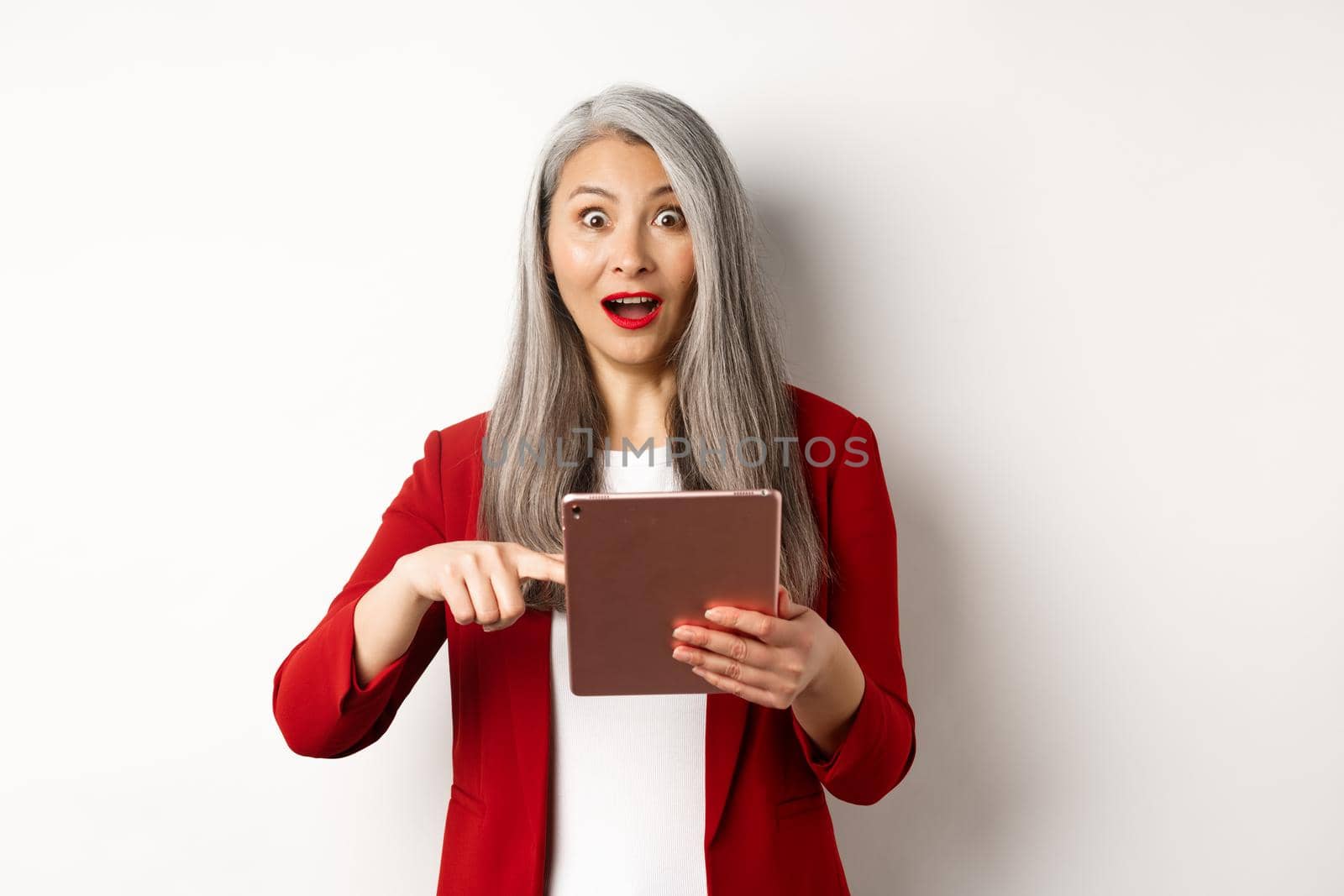 Business. Surprised female senior employer pointing at digital tablet screen, staring amazed at camera, checking out online promotion, white background by Benzoix