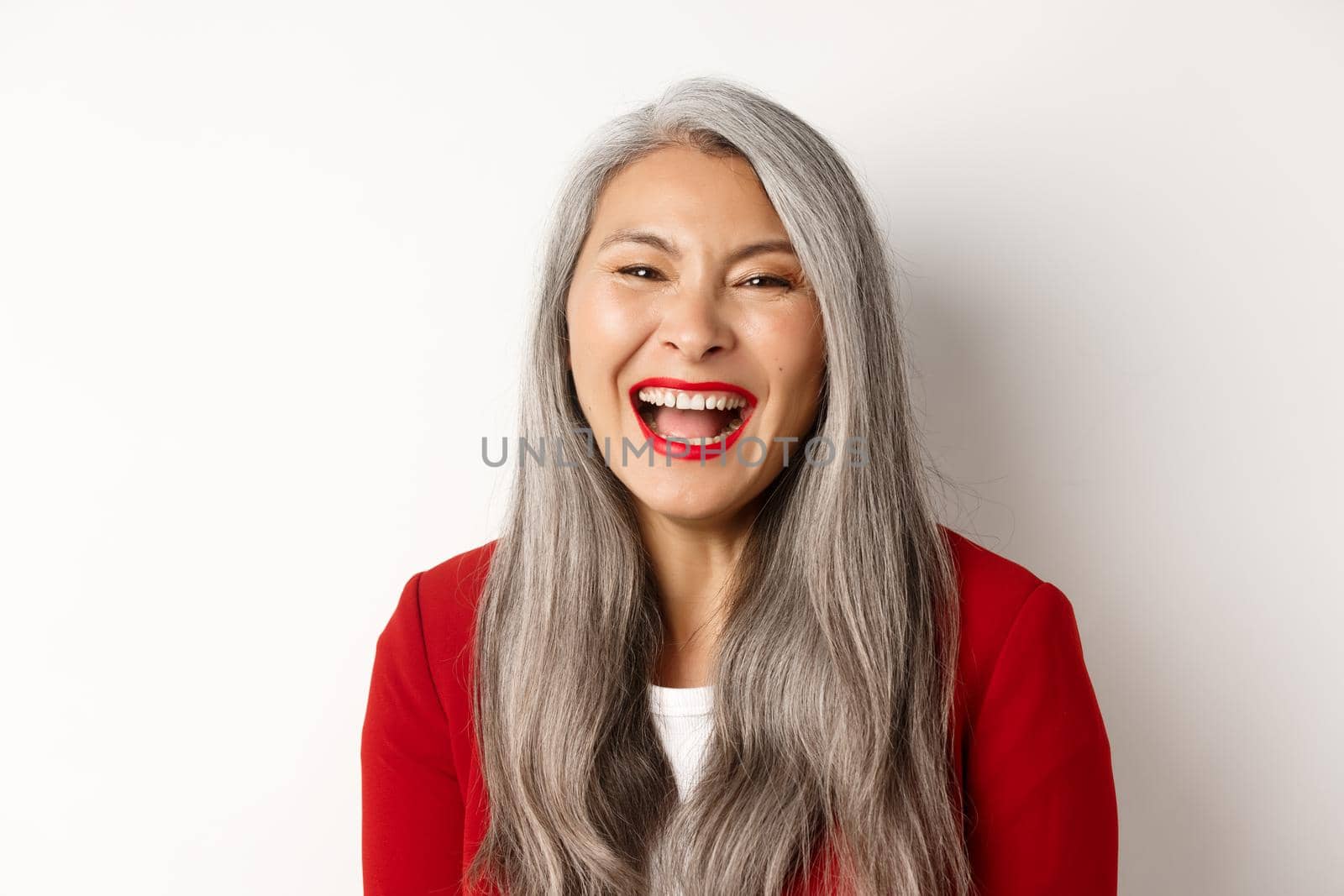 Close-up of happy asian businesswoman with long grey hair, wearing red blazer, laughing and smiling joyfully at camera, white background.