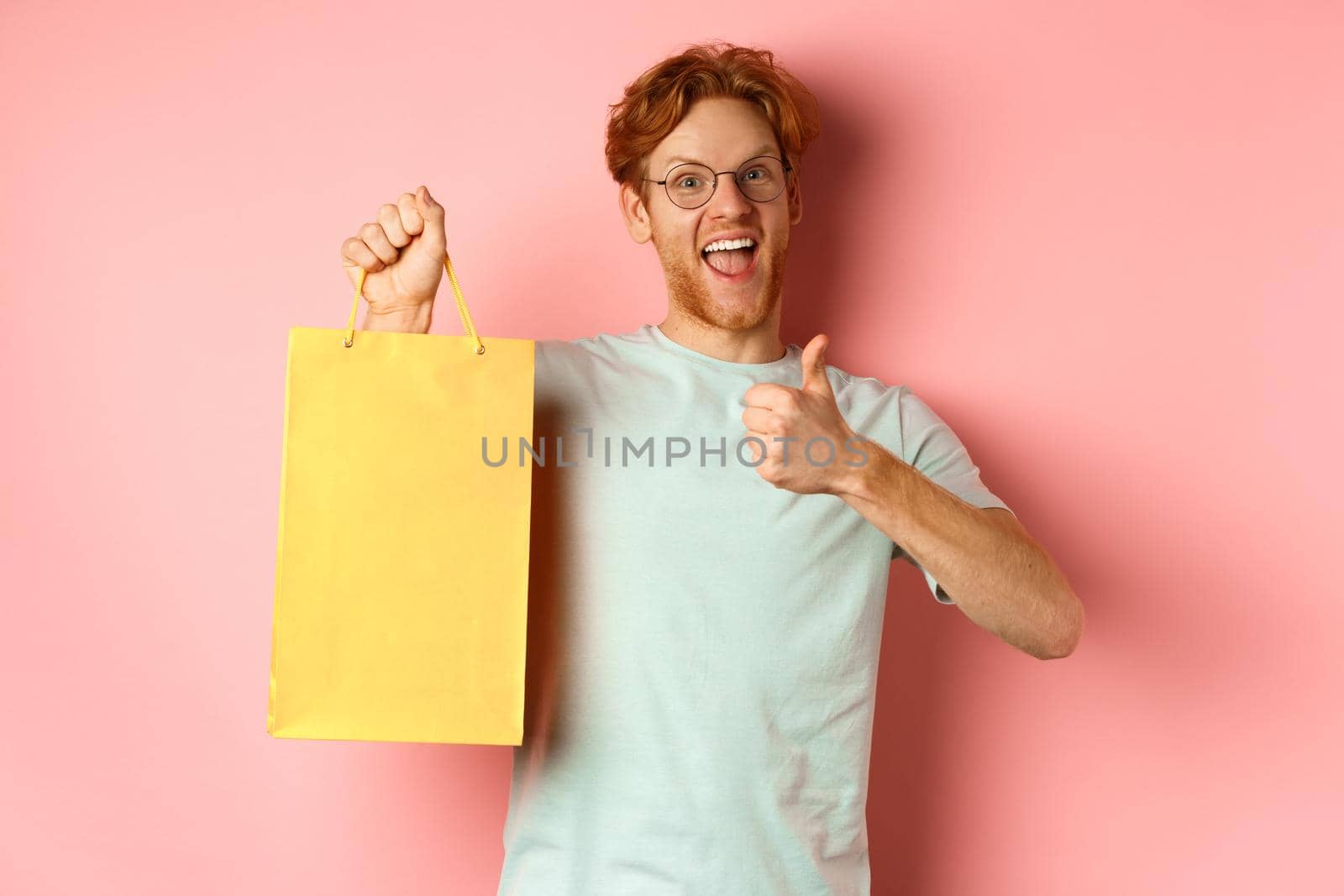 Cheerful redhead man in t-shirt and glasses pointing finger at shopping bag, showing shop with discounts, standing over pink background.