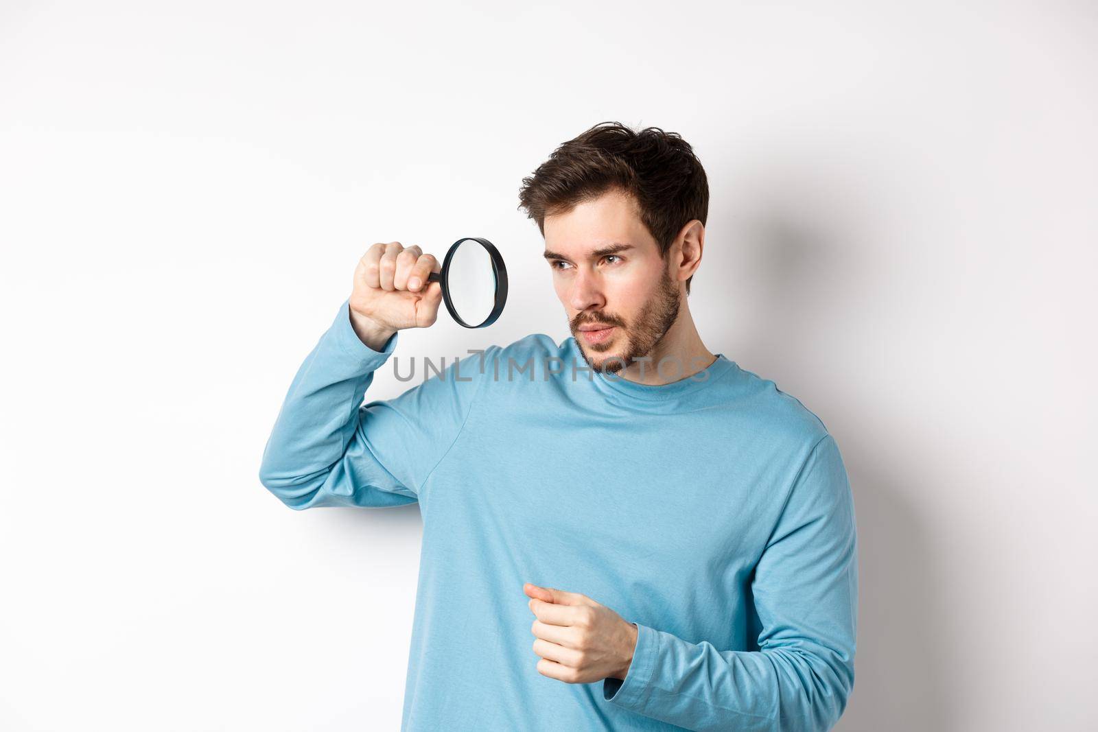 Young handsome man look through magnifying glass with curious face, investigating or searching for something, standing on white background by Benzoix