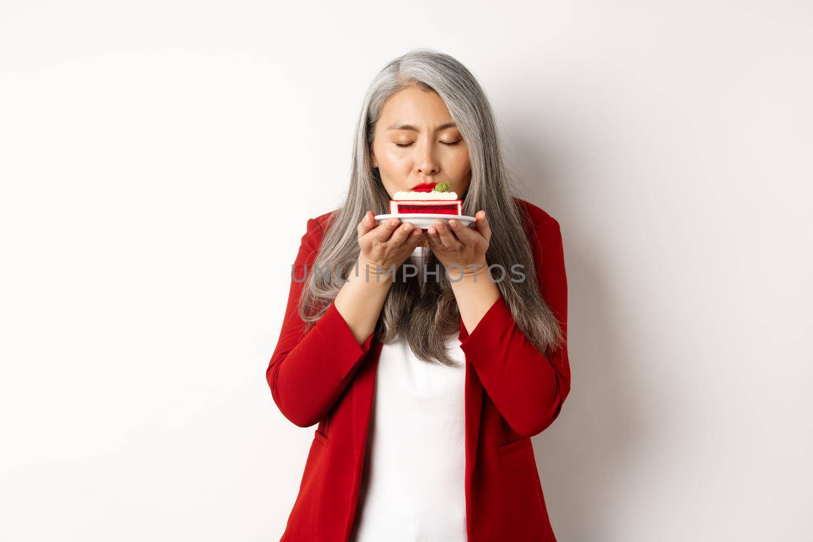 Image of funny asian senior lady sniffing delicious cake, smell dessert with satisfied face, standing over white background.