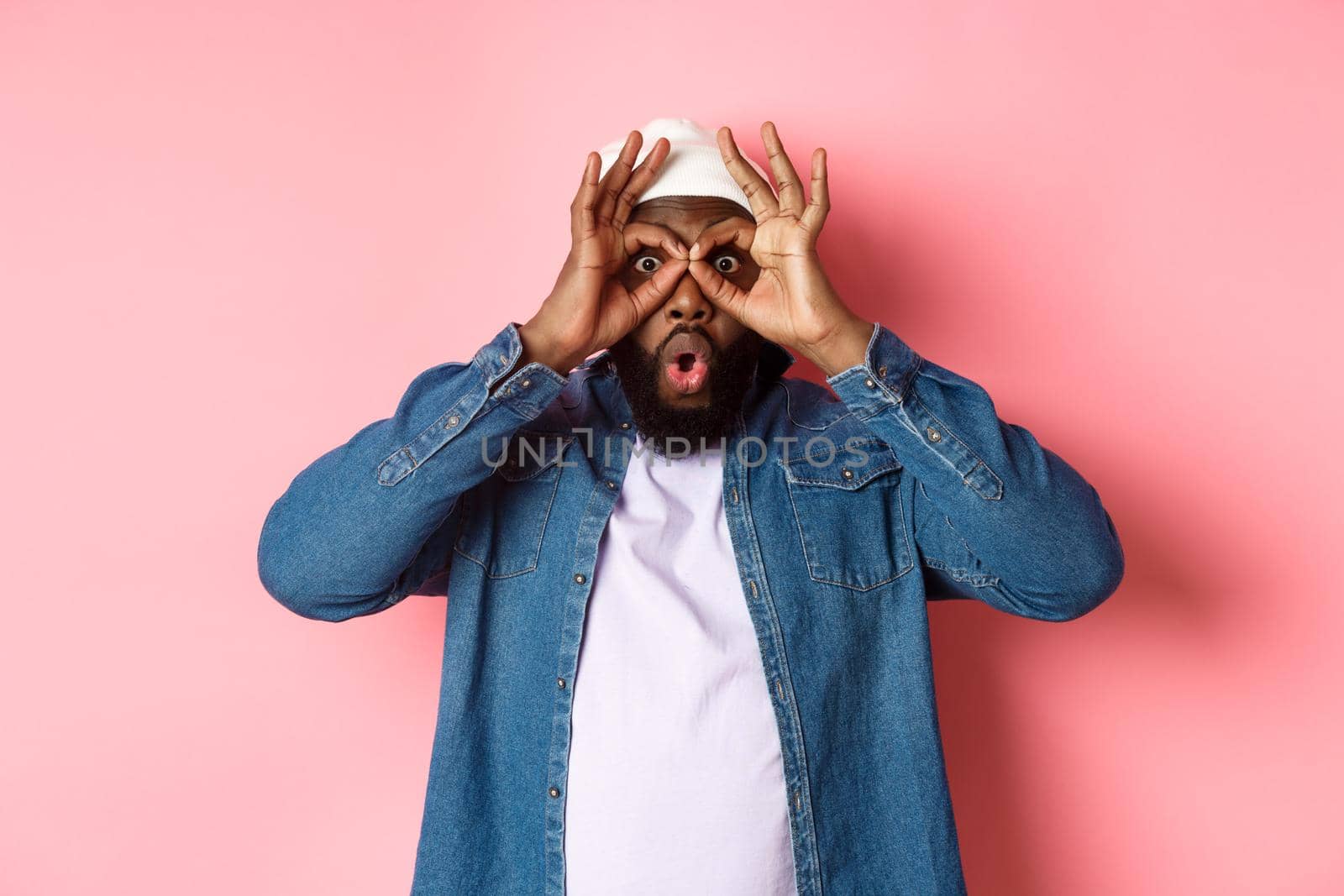 Shocked and impressed african-american guy staring through hand binoculars at camera, seeing amazing promo, standing over pink background by Benzoix