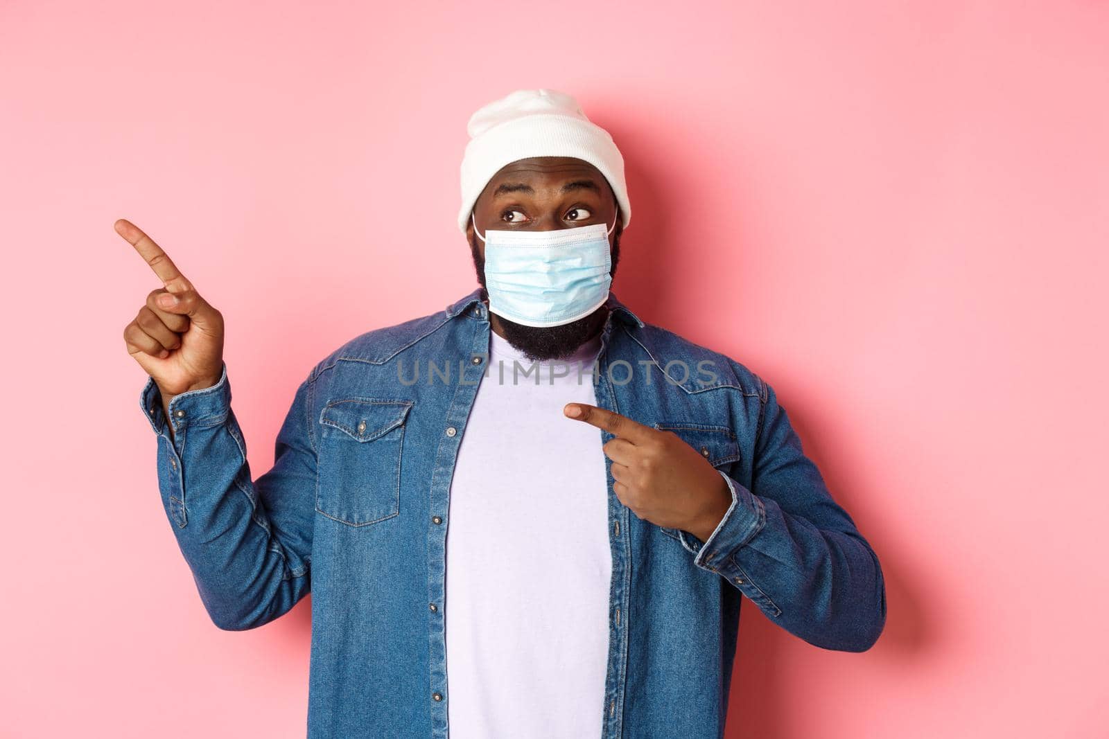 Coronavirus, lifestyle and global pandemic concept. Amused african-american male model in face mask pointing, looking left at promo offer, showing banner, pink background.