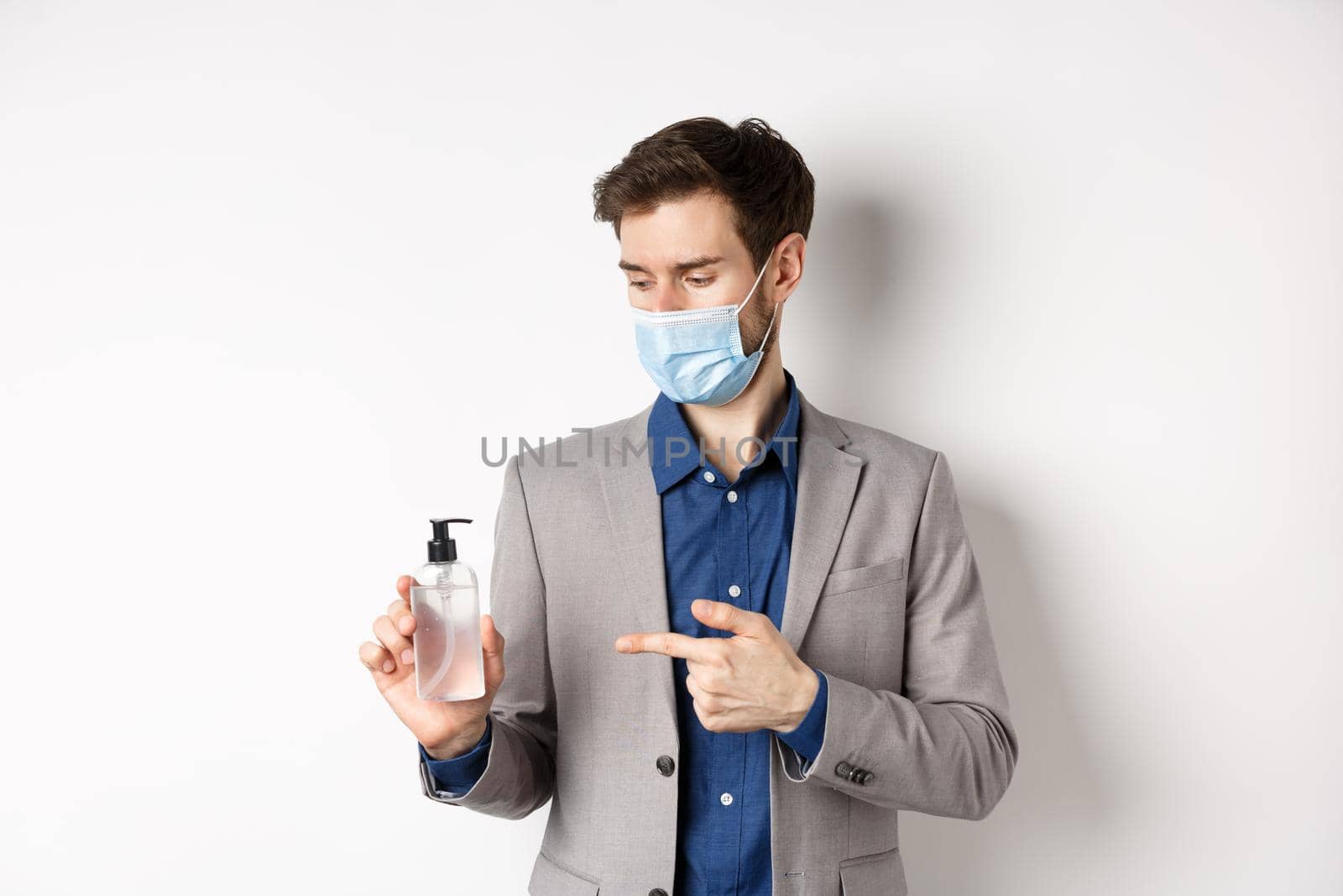 Covid-19, pandemic and business concept. Office worker in medical mask pointing at bottle of hand sanitizer, using antiseptic at work, white background by Benzoix