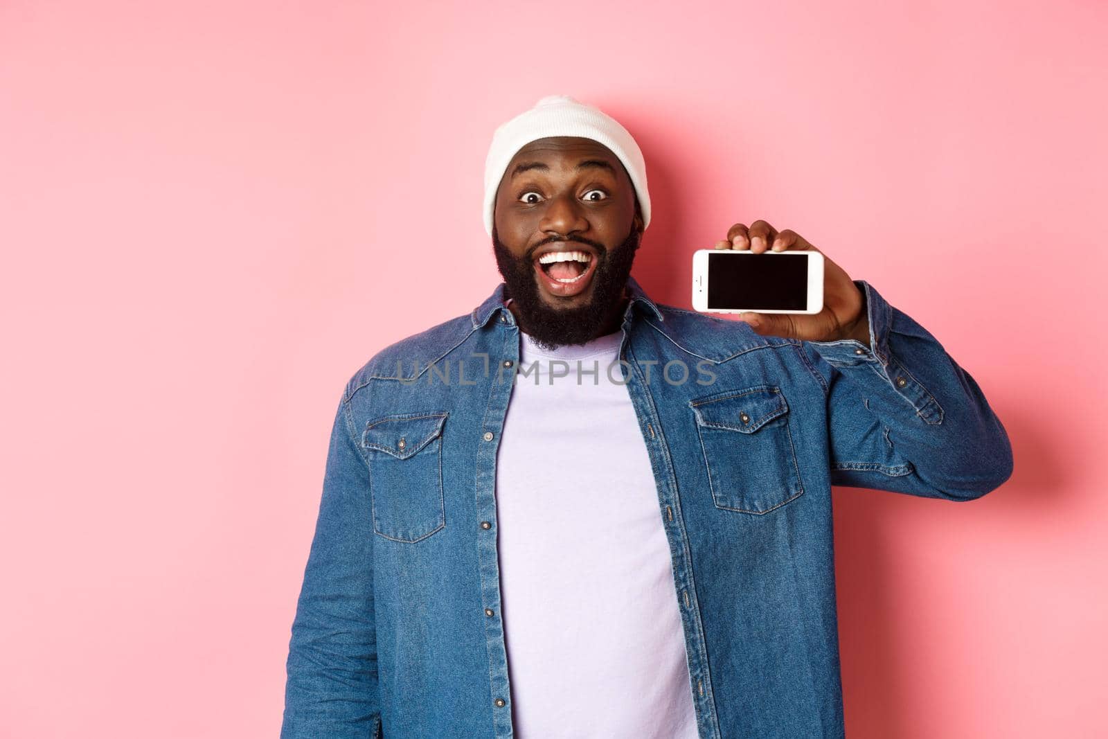 Online shopping and technology concept. Amazed Black man showing phone screen horizontally and staring at camera excited, standing over pink background by Benzoix