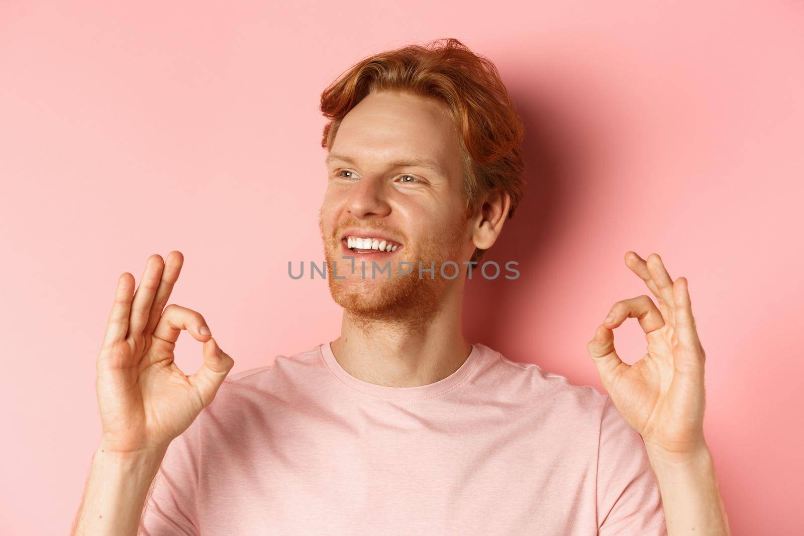 Close up of handsome redhead man in t-shirt, smiling satisfied and looking left, showing okay signs in approval, praising awesome product, standing over pink background by Benzoix