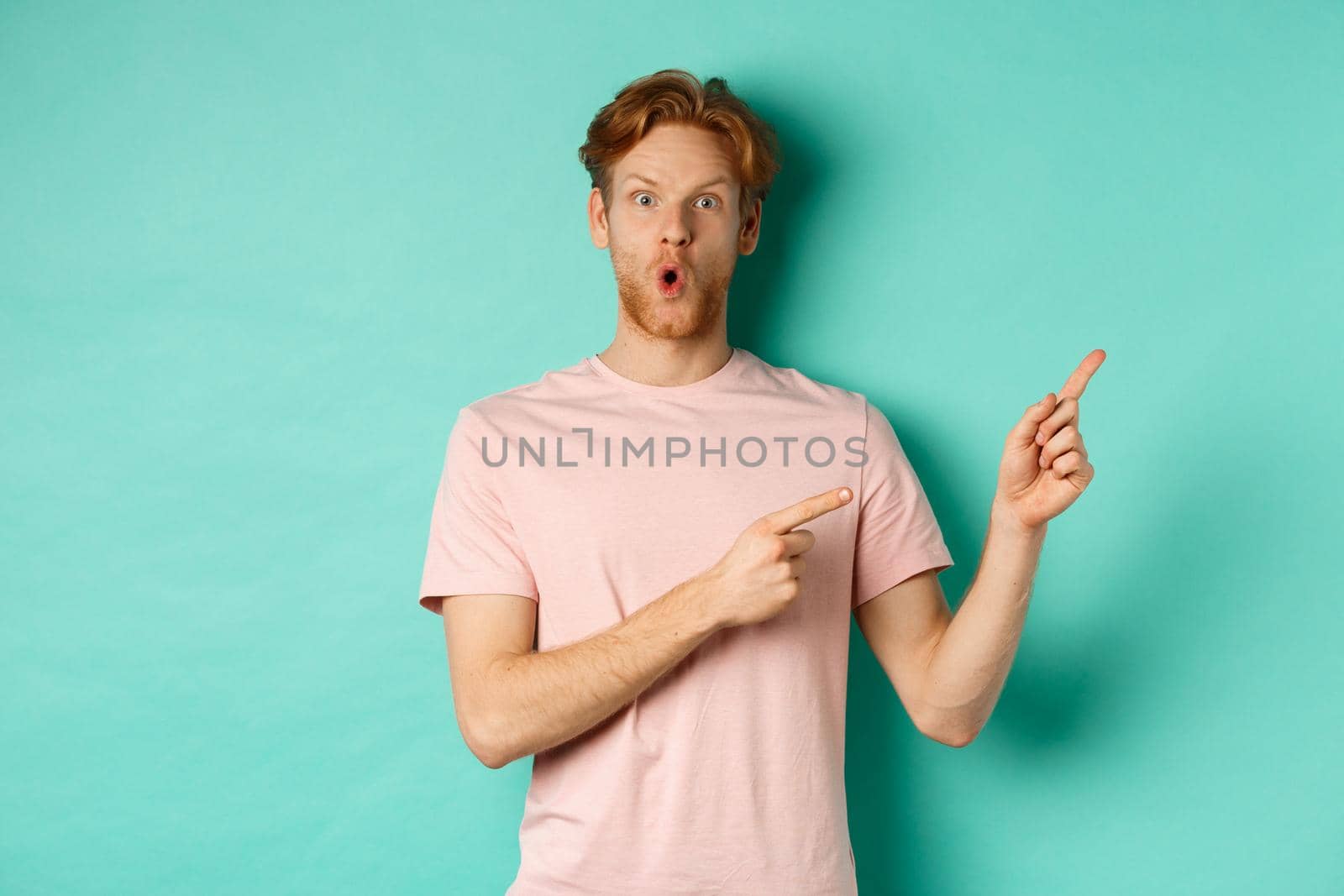 Handsome caucasian man with red hair, gasping in awe, pointing fingers at upper right corner, checking out promotion deal, standing over turquoise background.