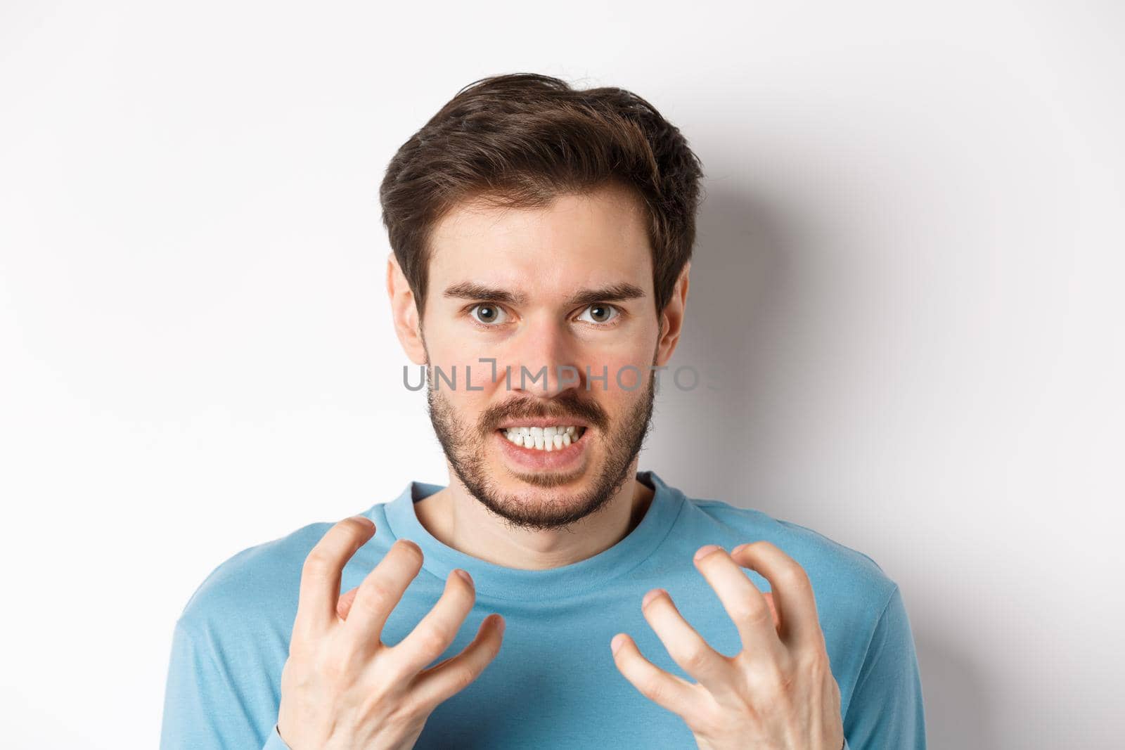 Close up of angry young man with beard, shaking hands mad, squeeze teeth and frowning furious, standing outraged over white background by Benzoix