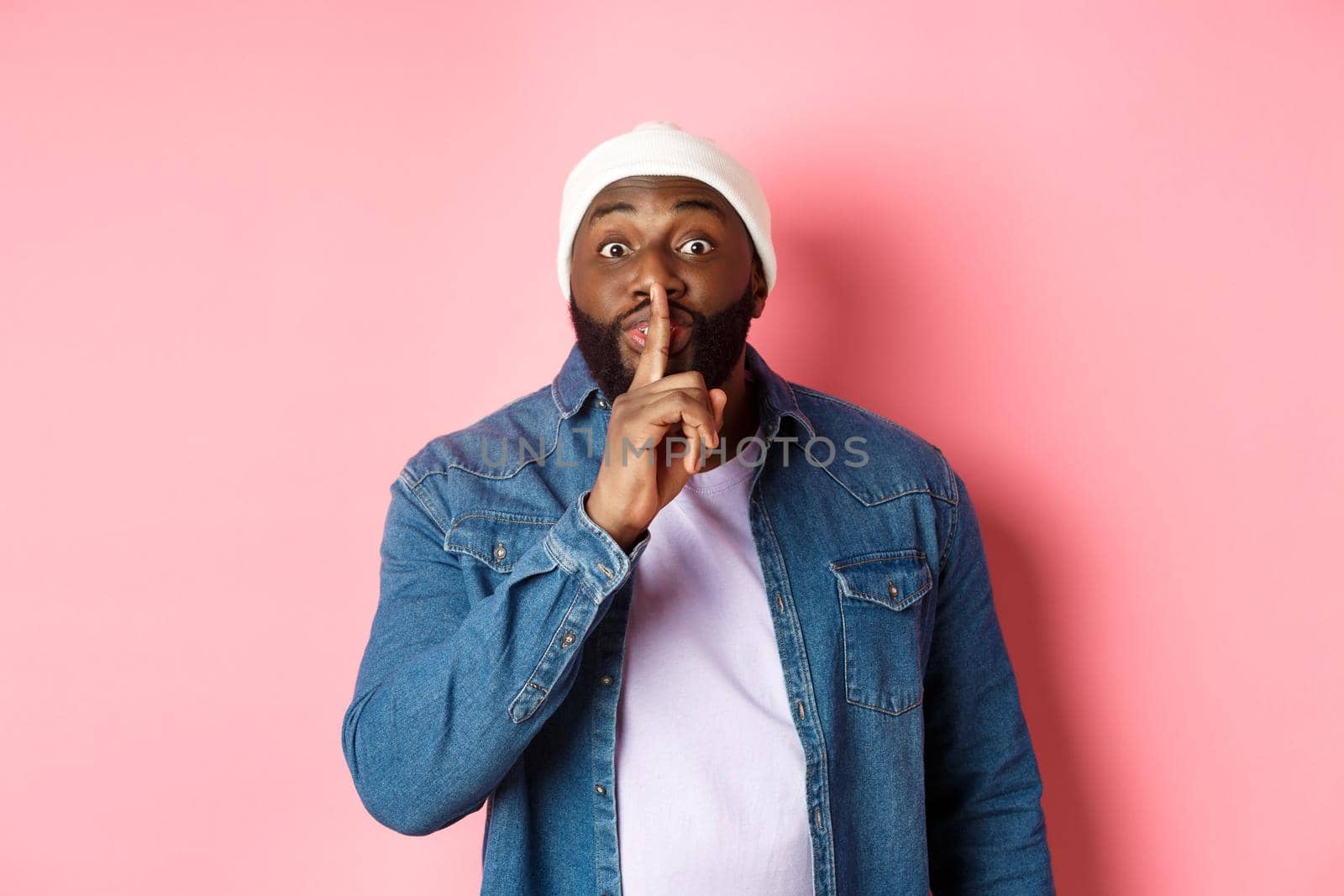 Handsome young black guy in beanie and denim shirt prepare surprise, hushing softly and looking at camera, asking keep quiet, pink background by Benzoix