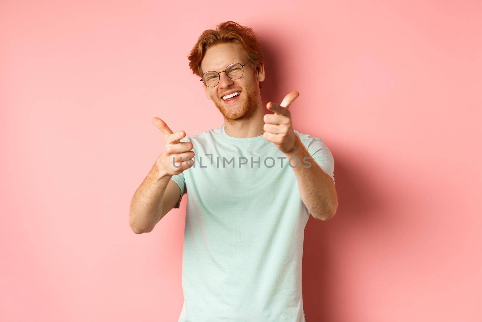 Happy young man with red hair and beard, wearing glasses, winking and smiling, pointing fingers at camera, choosing you, checking you something or congratulating winner.