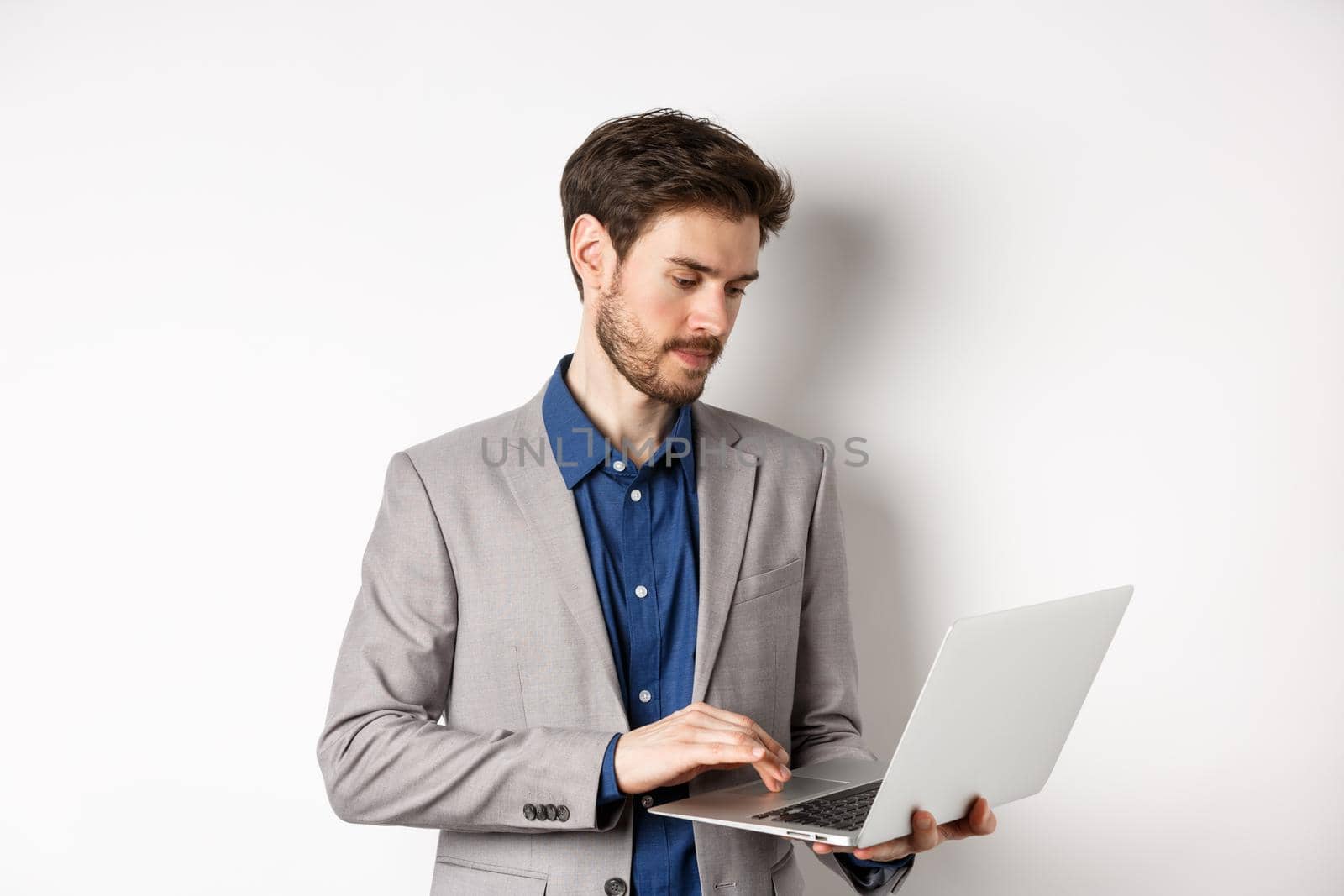 Handsome male entrepreneur working on laptop, looking serious at screen, standing against white background by Benzoix
