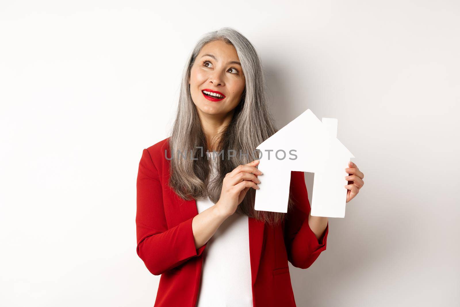 Dreamy senior woman thinking of buying property, showing paper house cutout and looking at upper left corner, standing over white background.