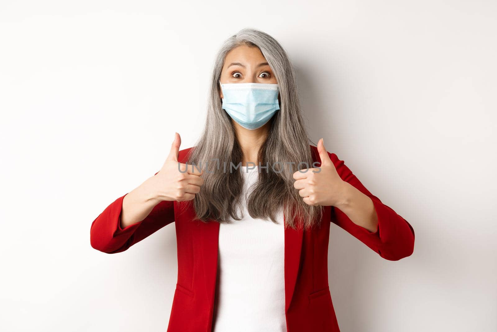 Coronavirus and business concept. Asian female manager in face mask looking cheerful, showing thumb-up in approval, white background.