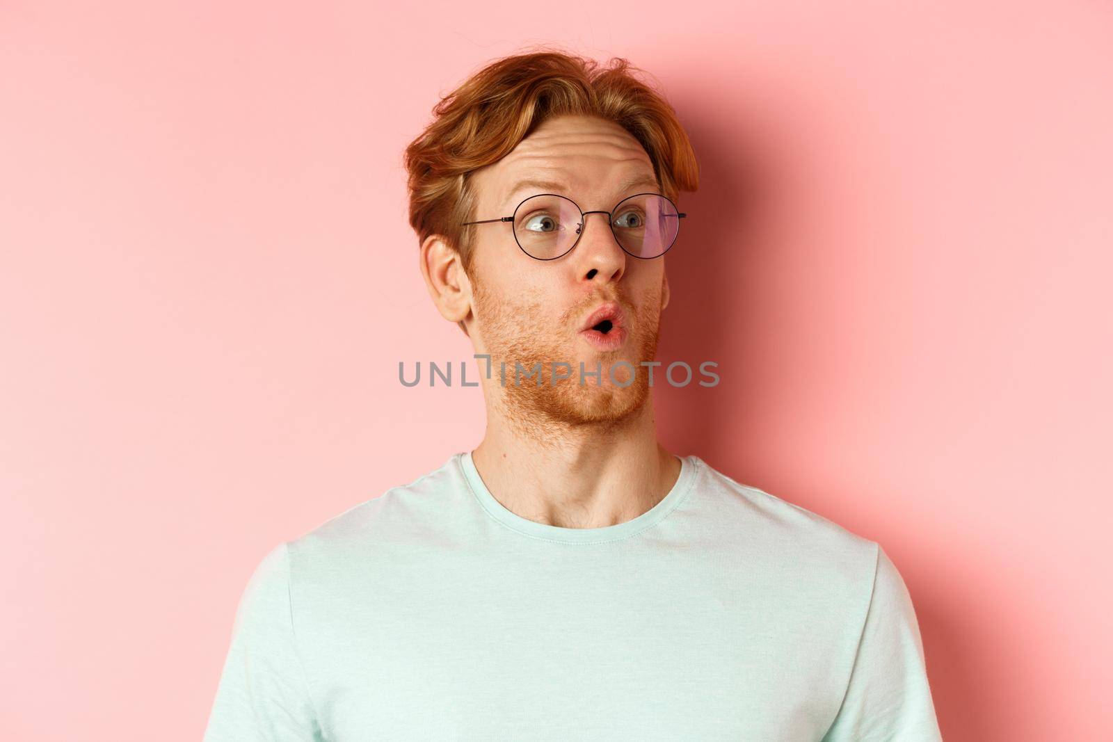 Face of intrigued young man with ginger hair and beard, raising eyebrows surpirsed and looking at upper right corner logo, standing over pink background.