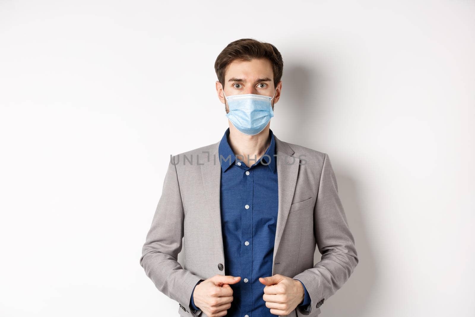 Covid-19 and office workers concept. Confident businessman in medical mask and suit looking at camera, standing on white background by Benzoix