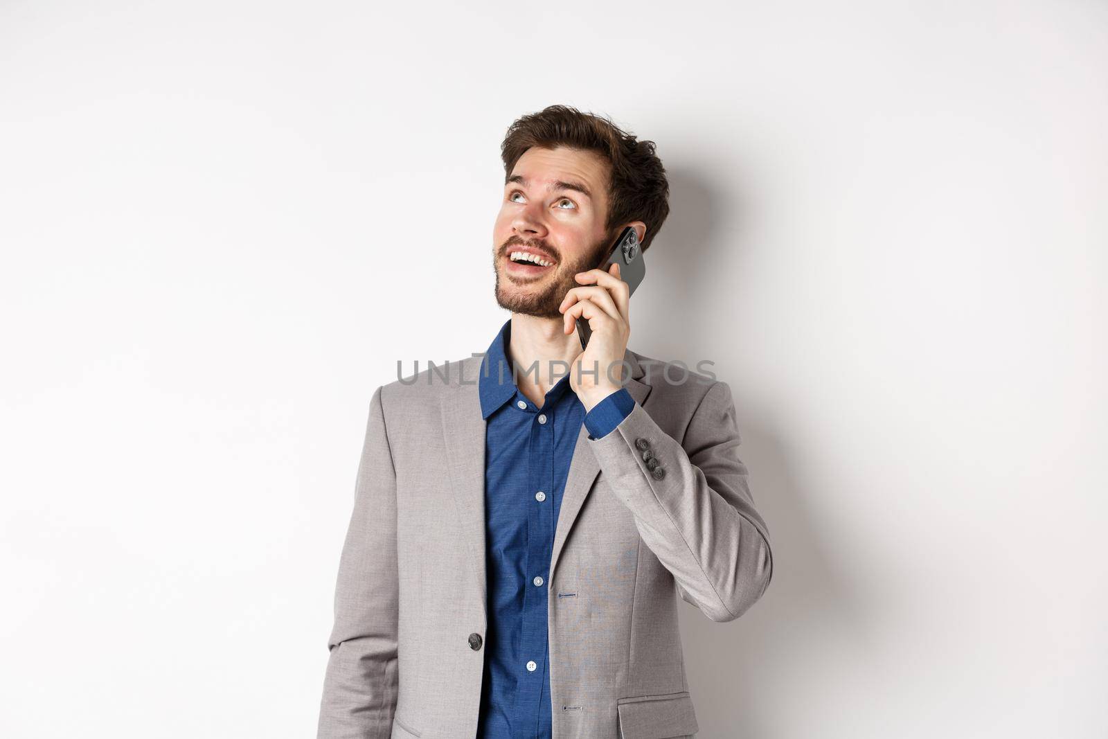 Carefree young man talking on mobile phone and smiling, looking up with dreamy face, standing in suit on white background.
