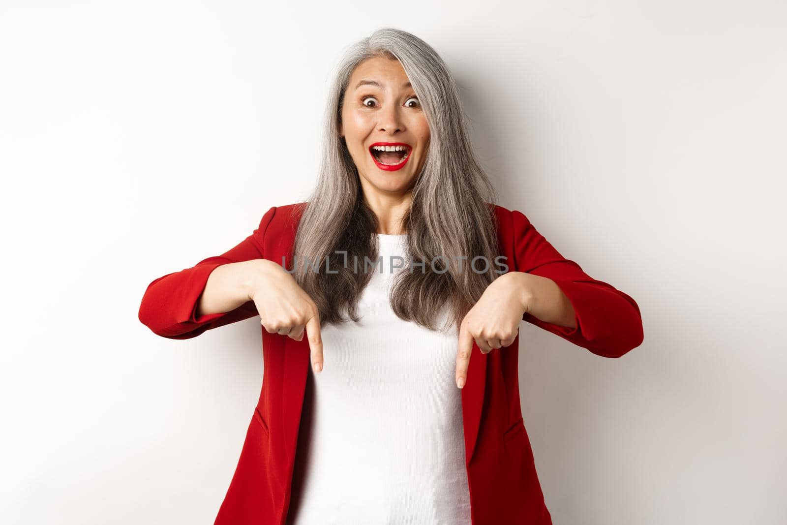 Portrait of happy asian lady in red blazer showing logo, pointing fingers down and smiling cheerful, check this out gesture, white background by Benzoix