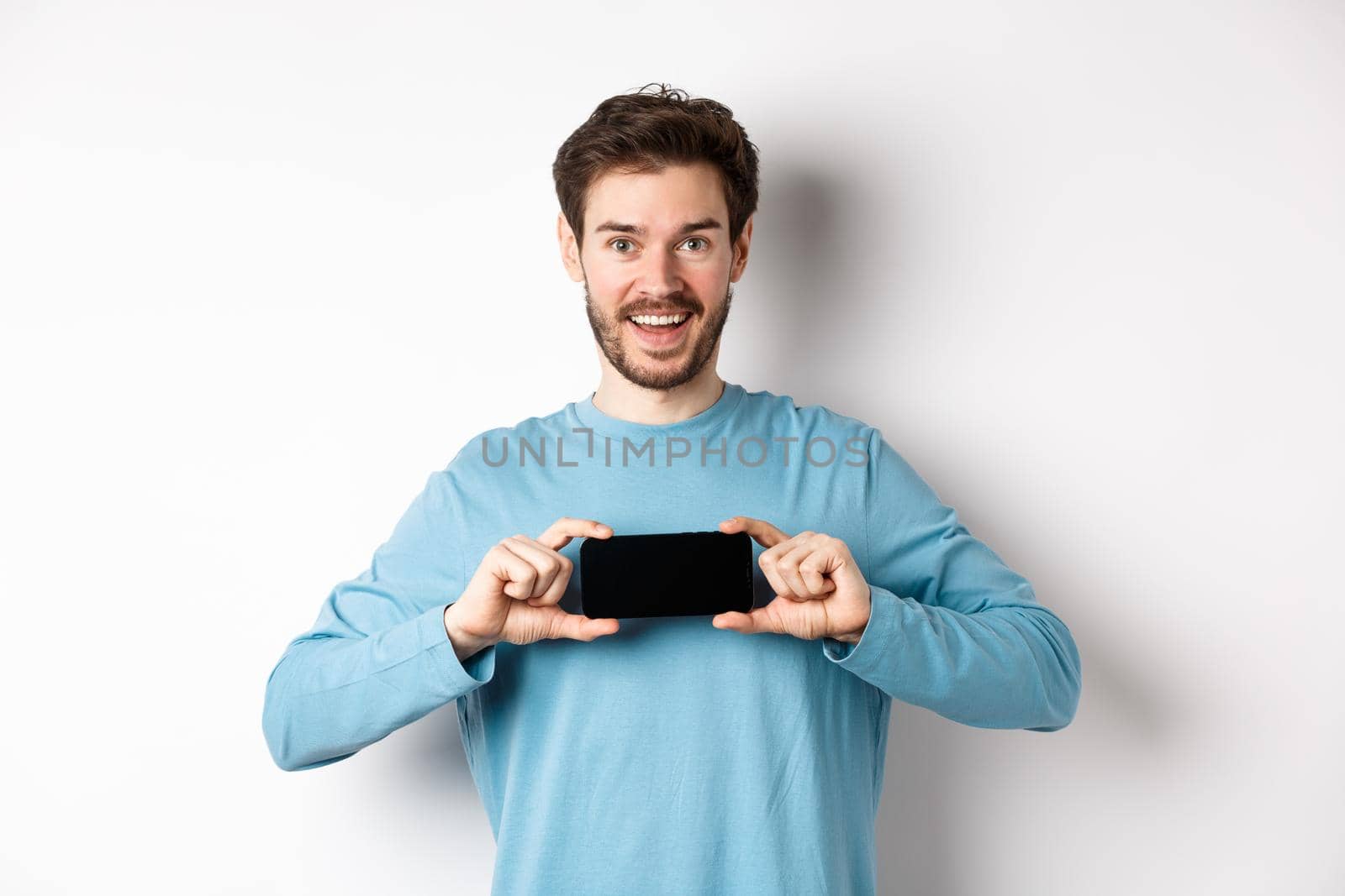 E-commerce and shopping concept. Smiling cheerful man showing empty smartphone screen in horizontal position, looking happy at camera, white background.