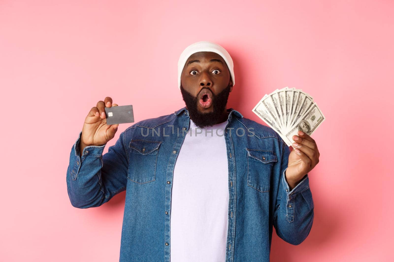 Shopping concept. Excited african-american man showing credit card and dollars, got deposit or money loan, standing over pink background.