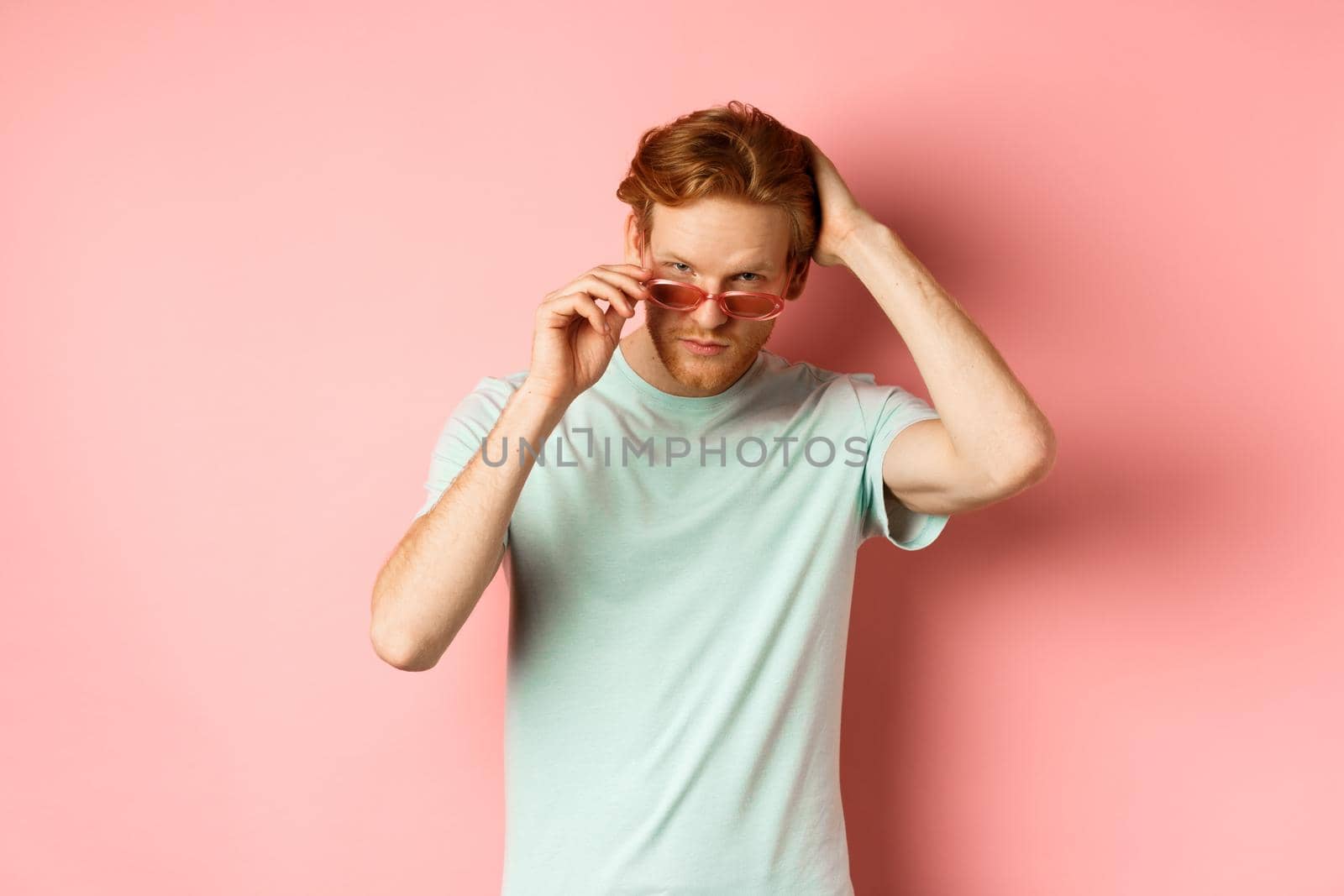 Handsome young redhead man in sunglasses, brushing hair with hand and looking smug and confident at camera, standing over pink background.