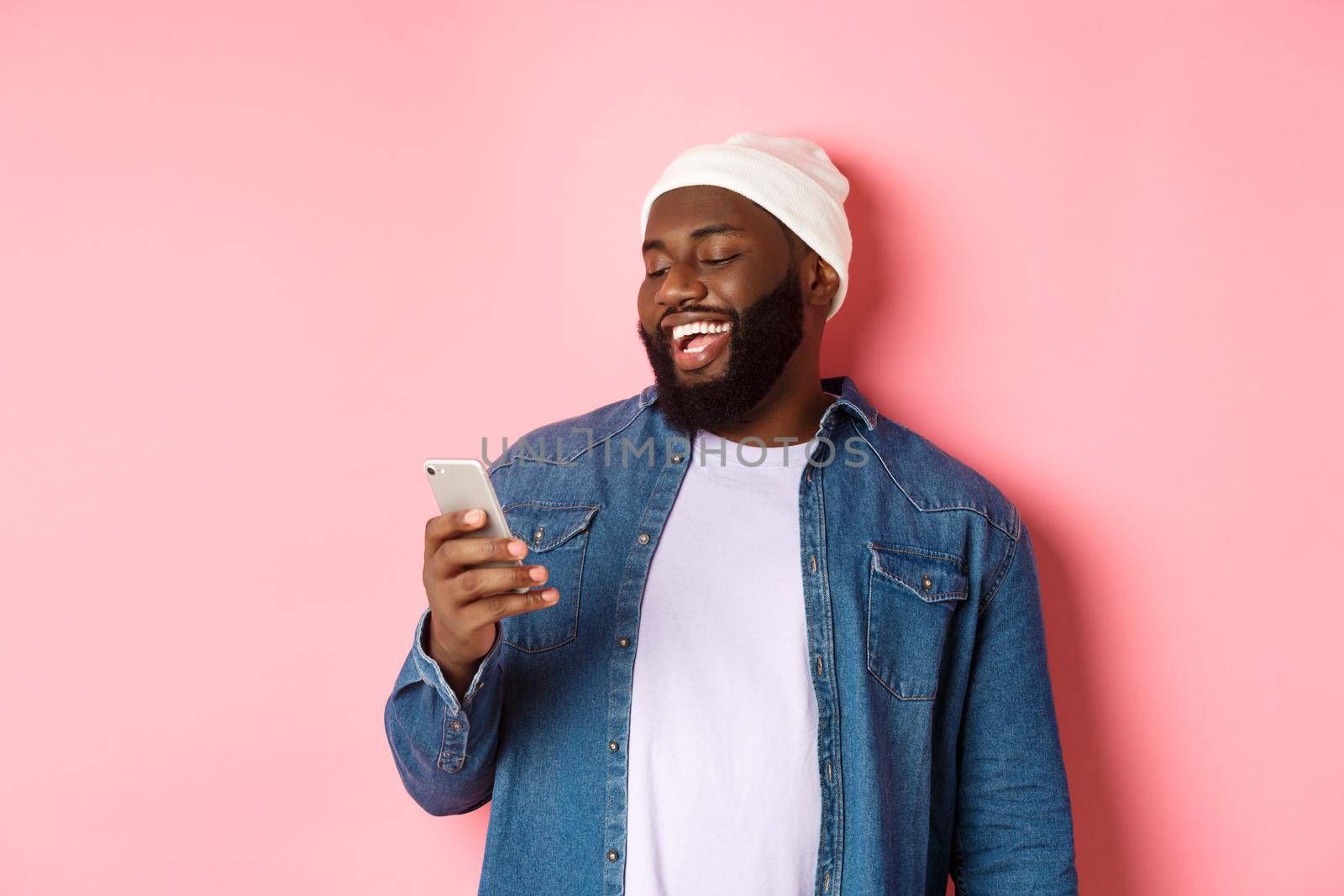 Technology and online shopping concept. Happy Black bearded man reading message and smiling, using smartphone against pink background.
