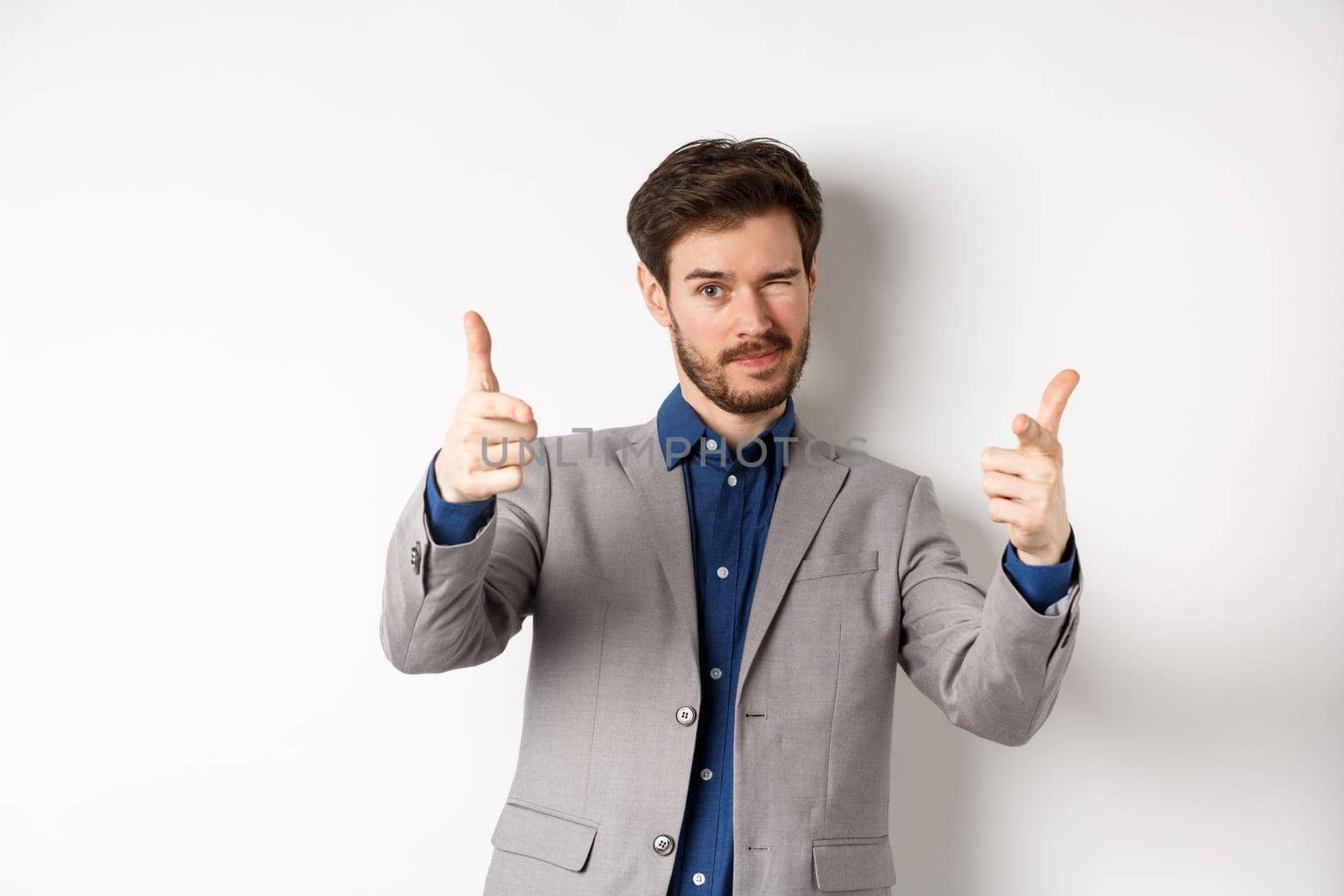 You got this. Handsome caucasian businessman in suit pointing fingers at camera and winking, motivating people, inviting to work in company, standing on white background.