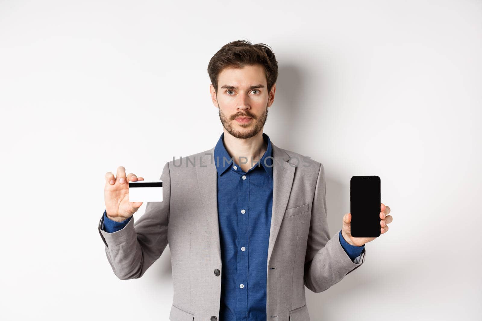 Online shopping. Serious bearded man in business suit showing plastic credit card with empty smartphone screen, standing against white background by Benzoix